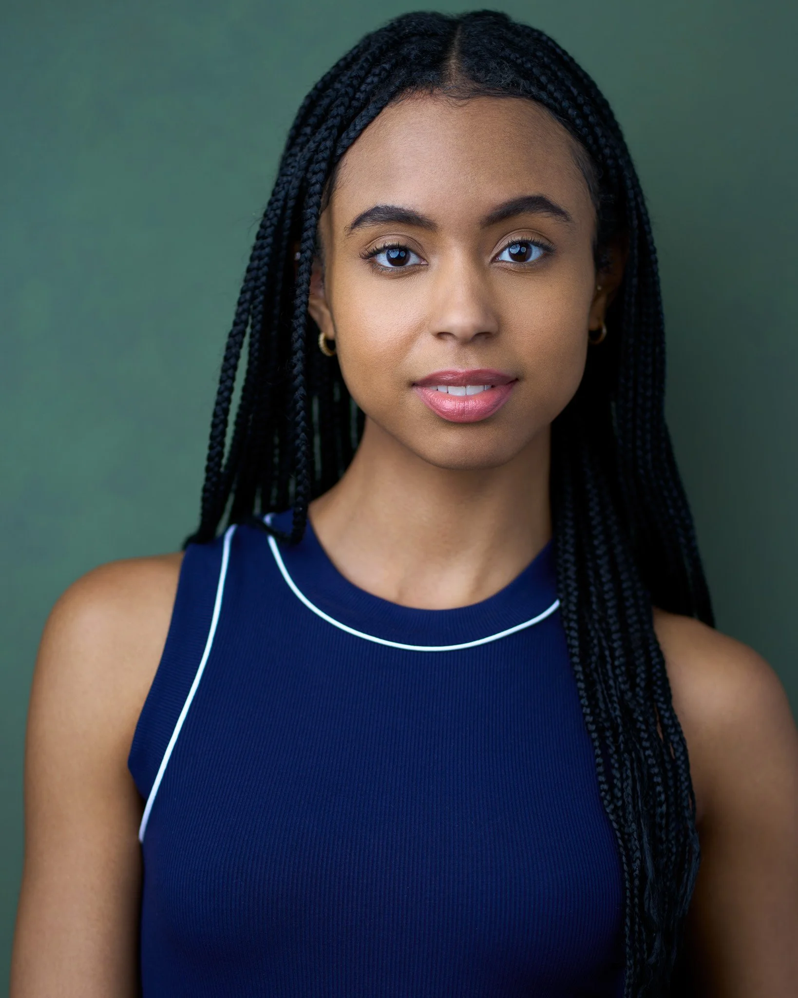 Acting headshot of a young woman with long braided hair, wearing a navy sleeveless top with white trim, against a green background, conveying a calm and confident expression.