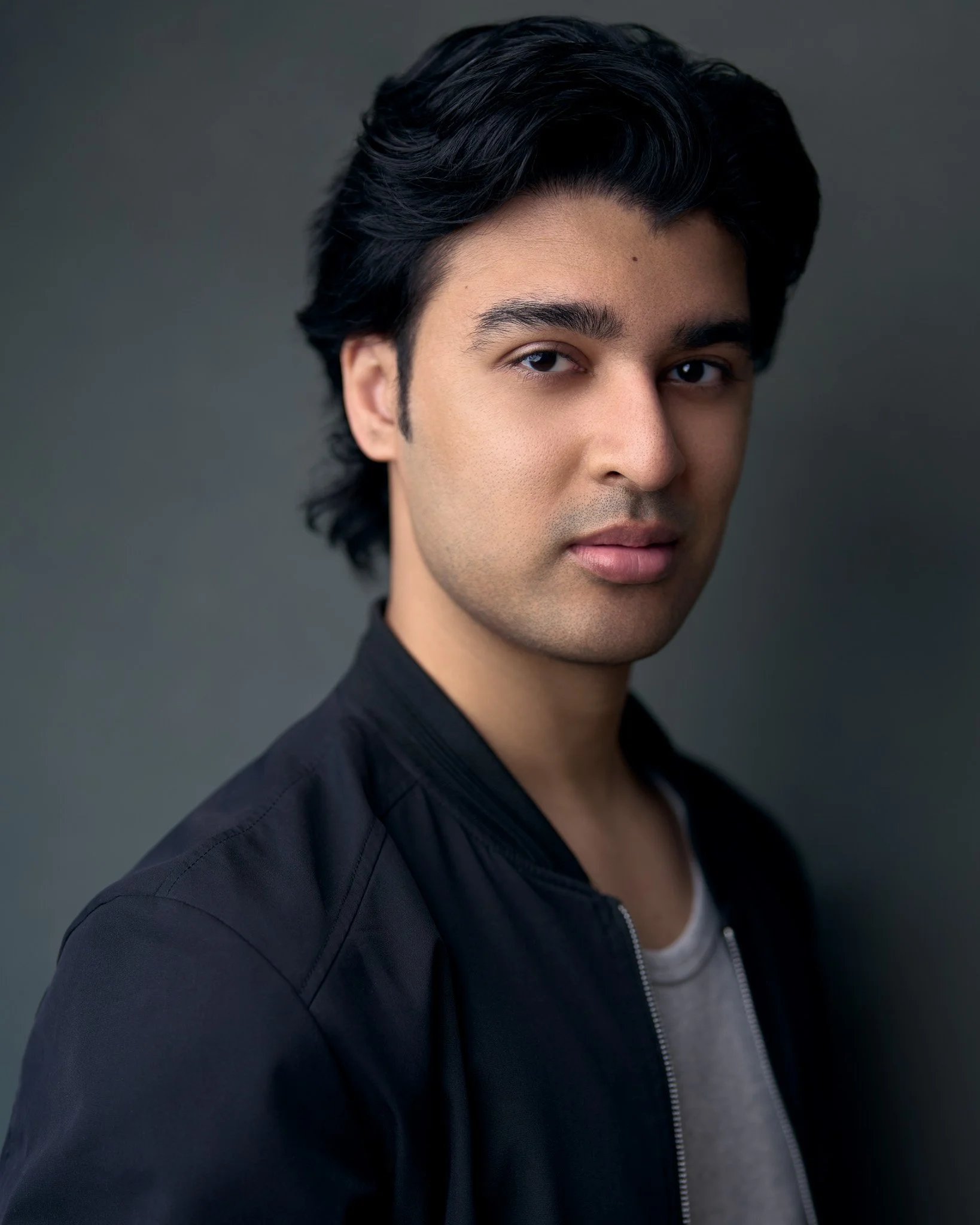 Acting headshot of a young man with dark hair and neutral expression wearing a black jacket over a gray shirt against a gray background, conveying a calm tone.