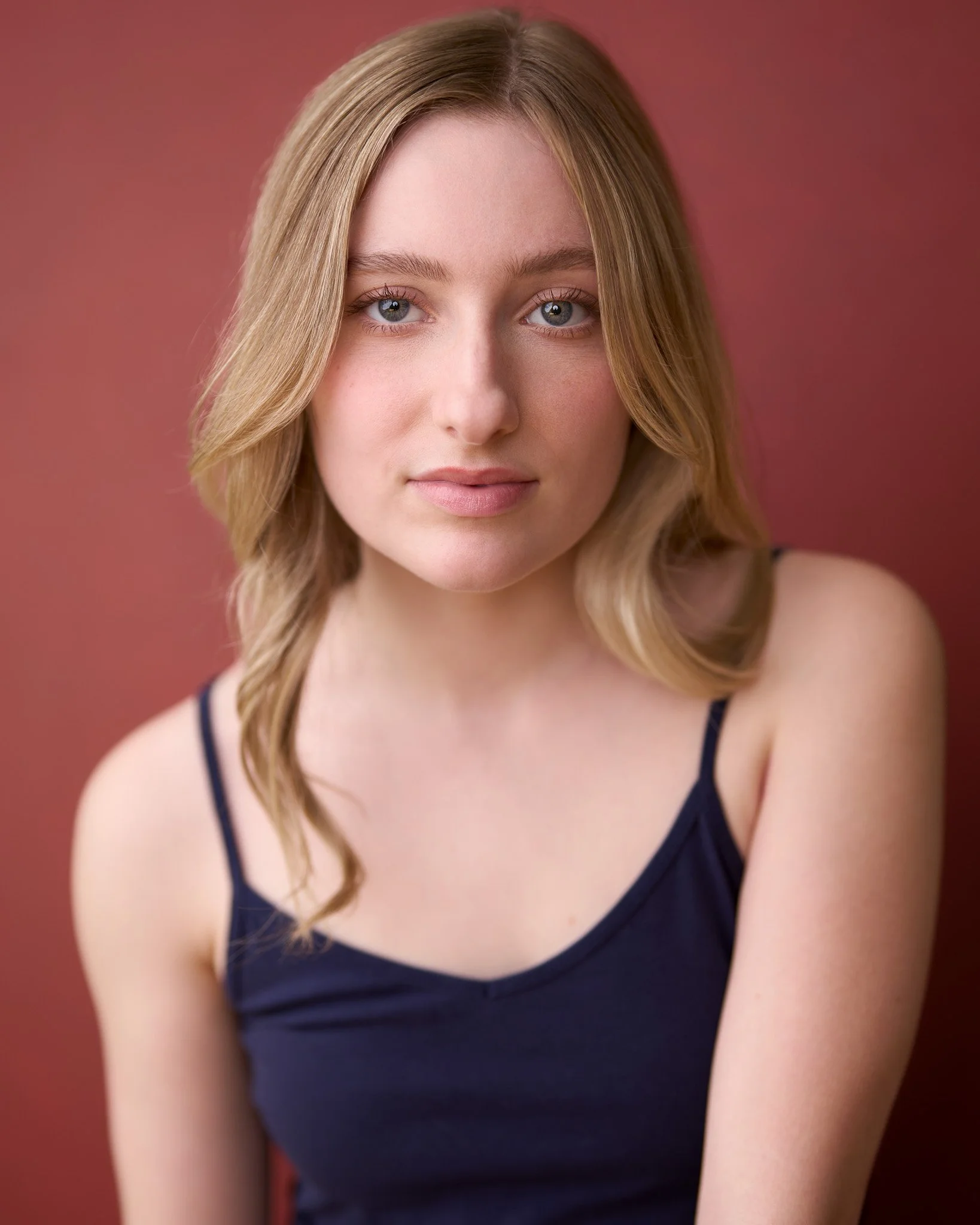 Close-up of a young woman with blonde hair, blue eyes, and light skin, wearing a dark blue top with thin straps, standing against a pink background.