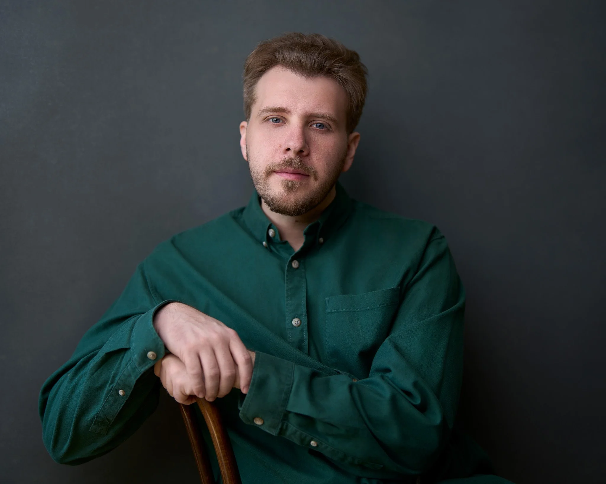 Acting headshot of a young man with brown hair and a beard, wearing a green button-up shirt, sitting with crossed arms against a gray background.