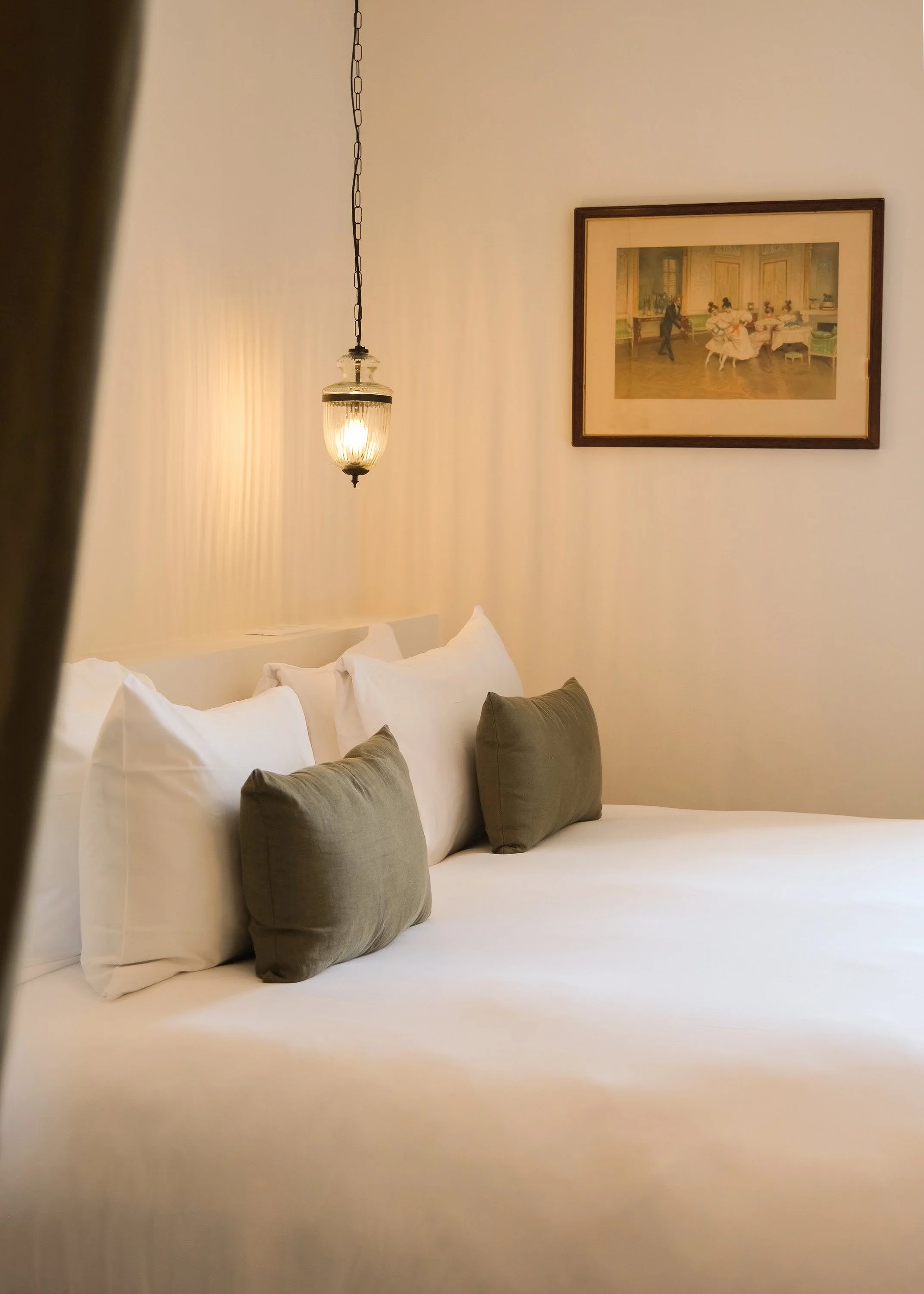 Lit de chambre avec coussins verts, lampe suspendue, tableau ancien au mur.