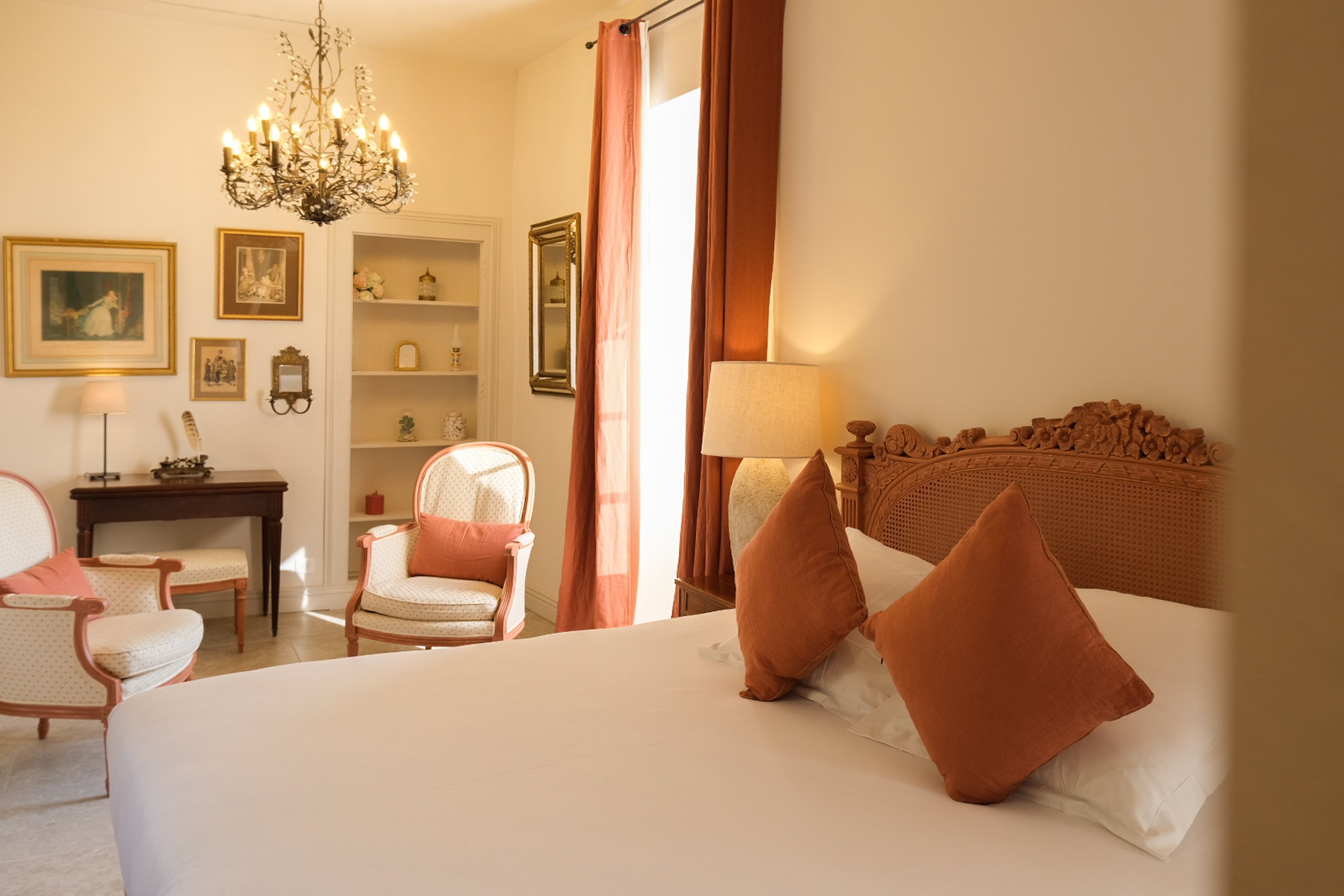 Chambre élégante avec un grand lit en bois sculpté, deux coussins marron, une lampe de chevet, une fenêtre avec rideaux oranges, deux fauteuils avec coussins, un chandelier suspendu au plafond, plusieurs œuvres d'art sur les murs, une étagère blanche