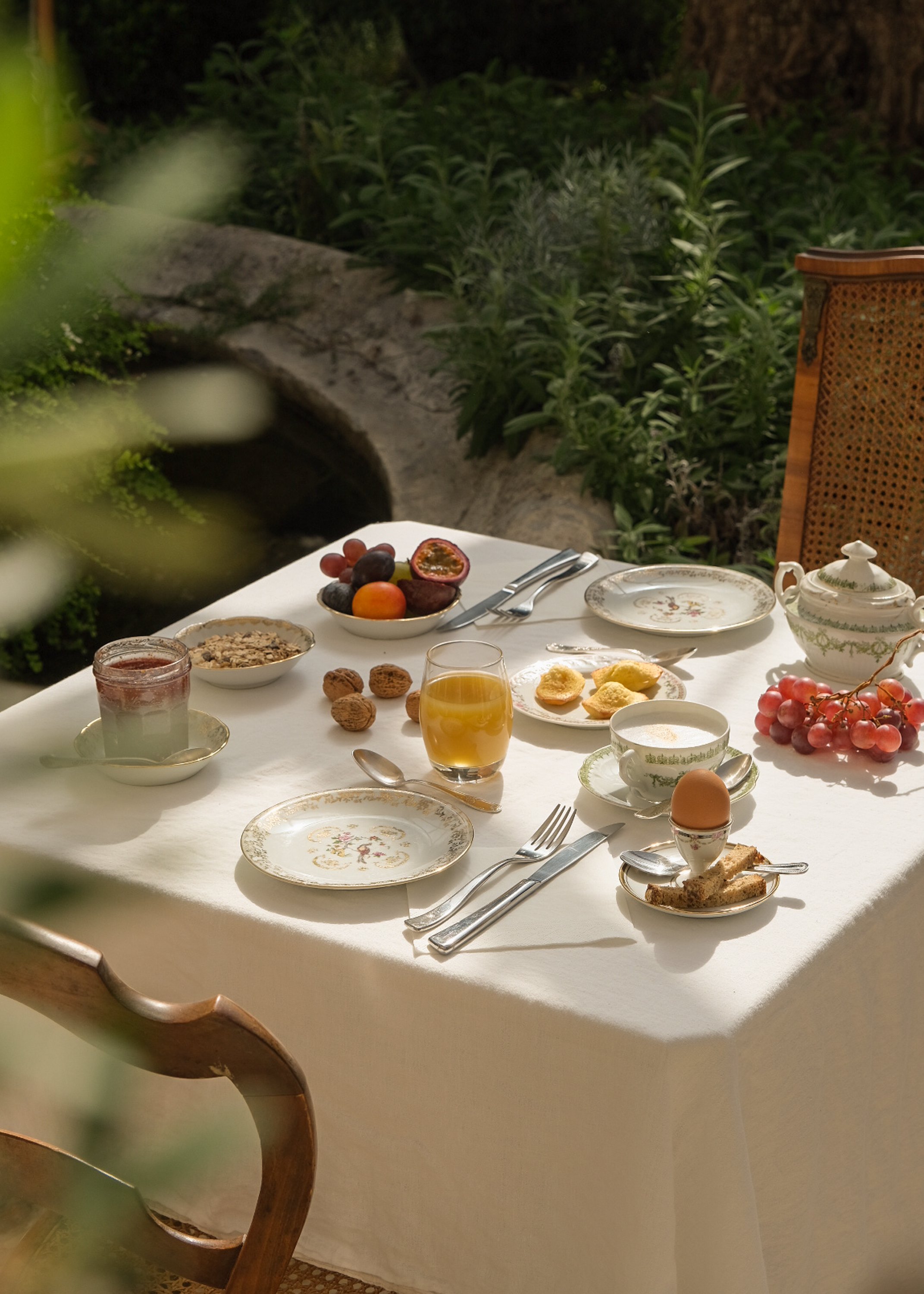 Petit déjeuner servi sur une table en extérieur, comprenant des fruits, un œuf, un verre de jus d'orange, du fromage blanc, des céréales, du pain, une cuillère, une fourchette, une tasse, des noix, et un pot de confiture, avec un décor naturel en arr