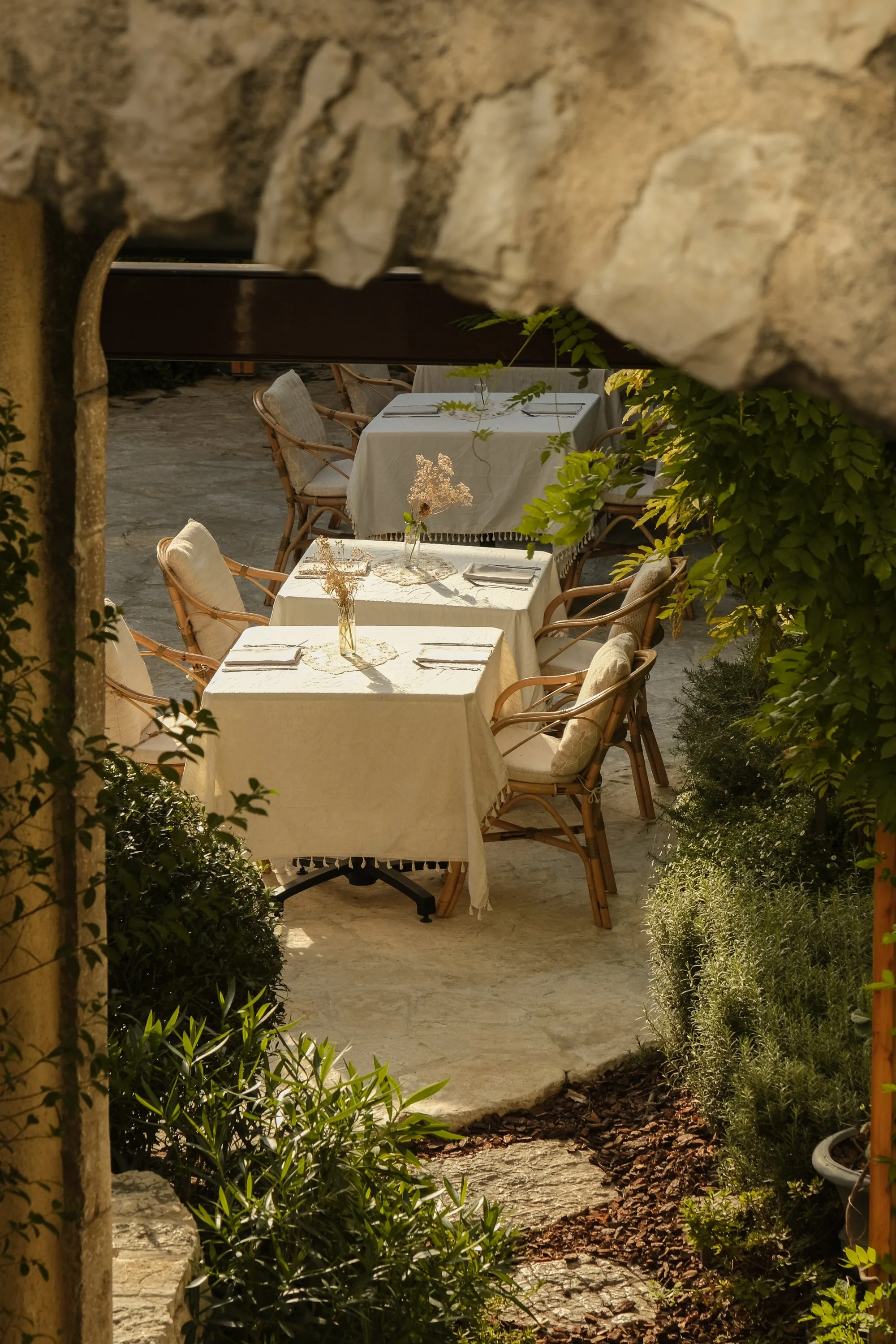 Deux tables de restaurant avec nappe blanche, décorées de vases avec des fleurs séchées, dans un jardin extérieur entouré de verdure, vue à travers une ouverture rocheuse.