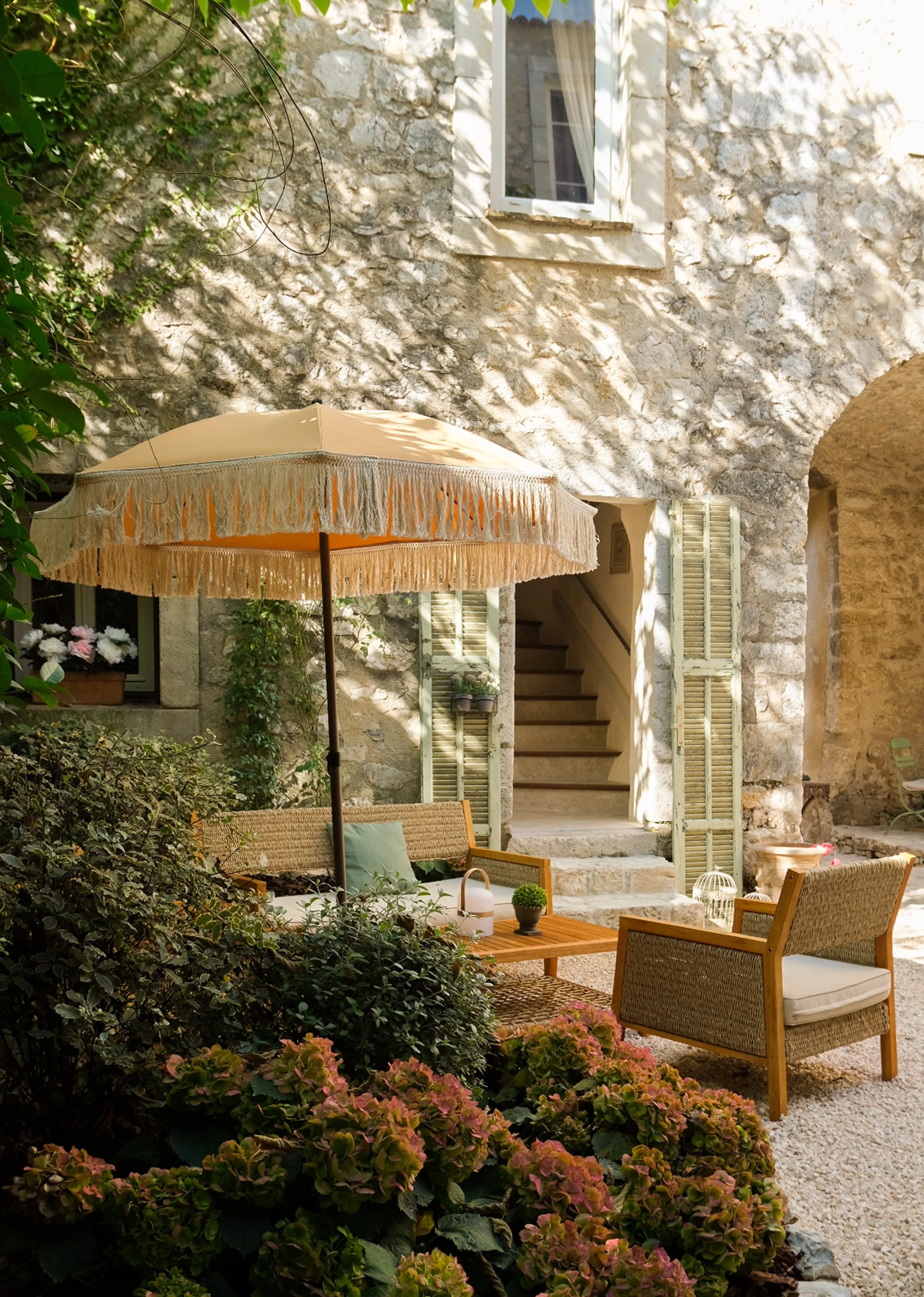 Un patio charmant avec un parasol beige, un canapé en rotin avec coussins, une table en bois, et des chaises, entourés de végétation et de fleurs, avec une maison en pierre en arrière-plan.