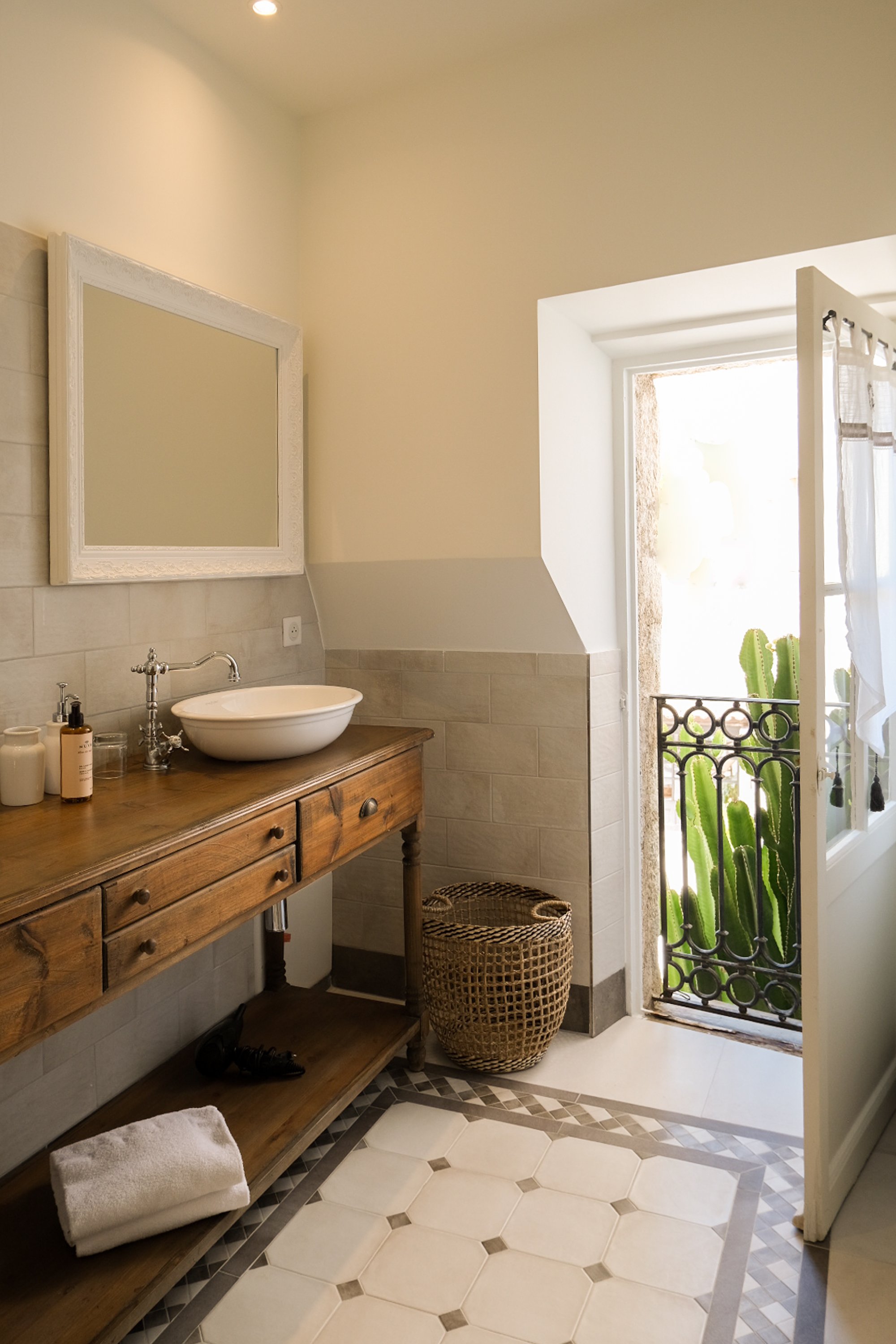 Une salle de bain lumineuse avec un lavabo sur une commode en bois, un miroir blanc, un panier en osier, et une porte donnant sur un balcon avec des cactus.