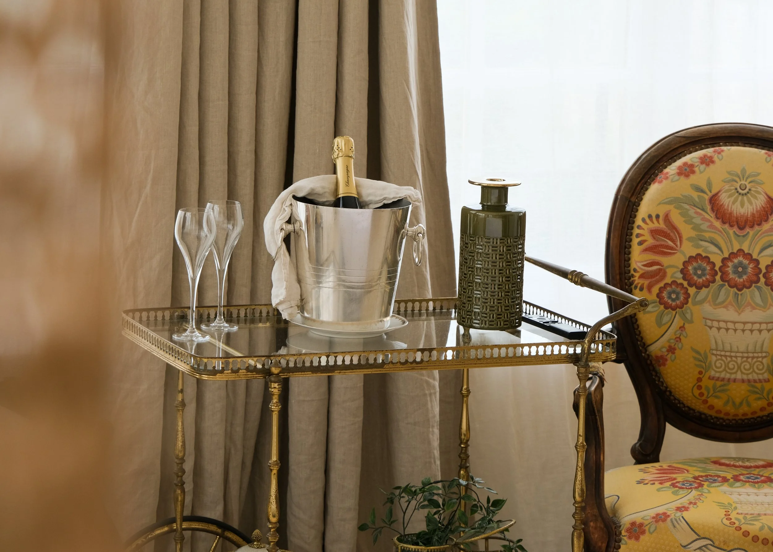 Une table en métal doré avec une bouteille de champagne dans un seau à glace, deux flûtes à champagne, une bouteille de vin ou de spiritueux et un stylo. À côté, une chaise en bois avec un rembourrage floral.