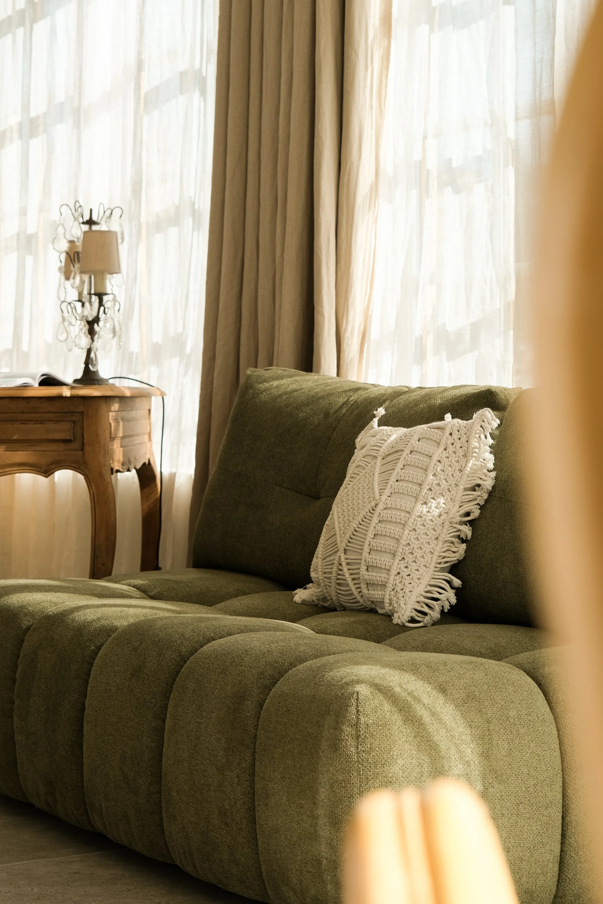 Un coussin en tricot blanc posé sur un canapé vert en tissu dans un salon lumineux, avec des rideaux beige clair derrière.