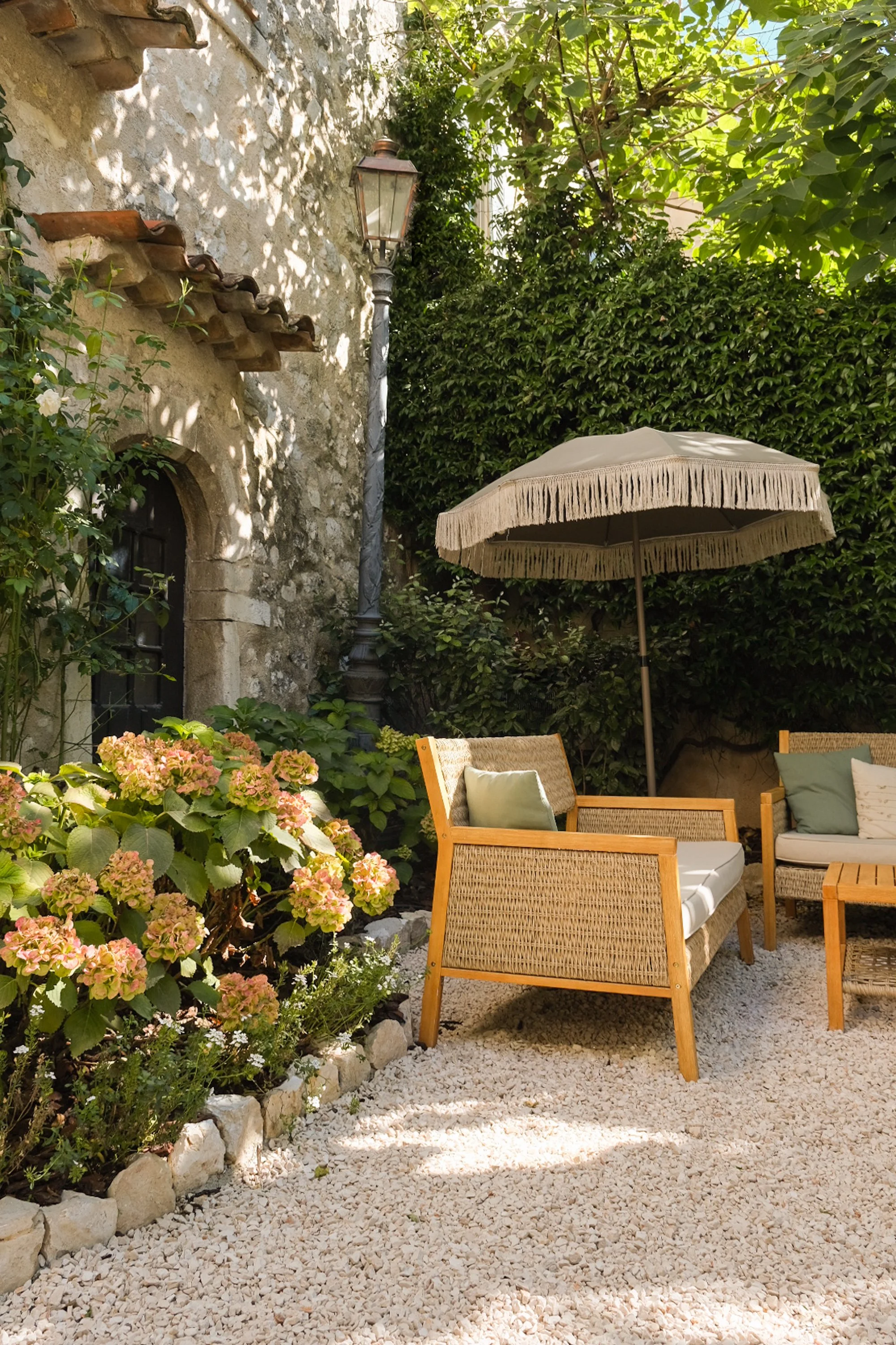 Une terrasse de jardin avec des coussins, un parasol et des fleurs, entourée de végétation et d'une vieille maison en pierre.