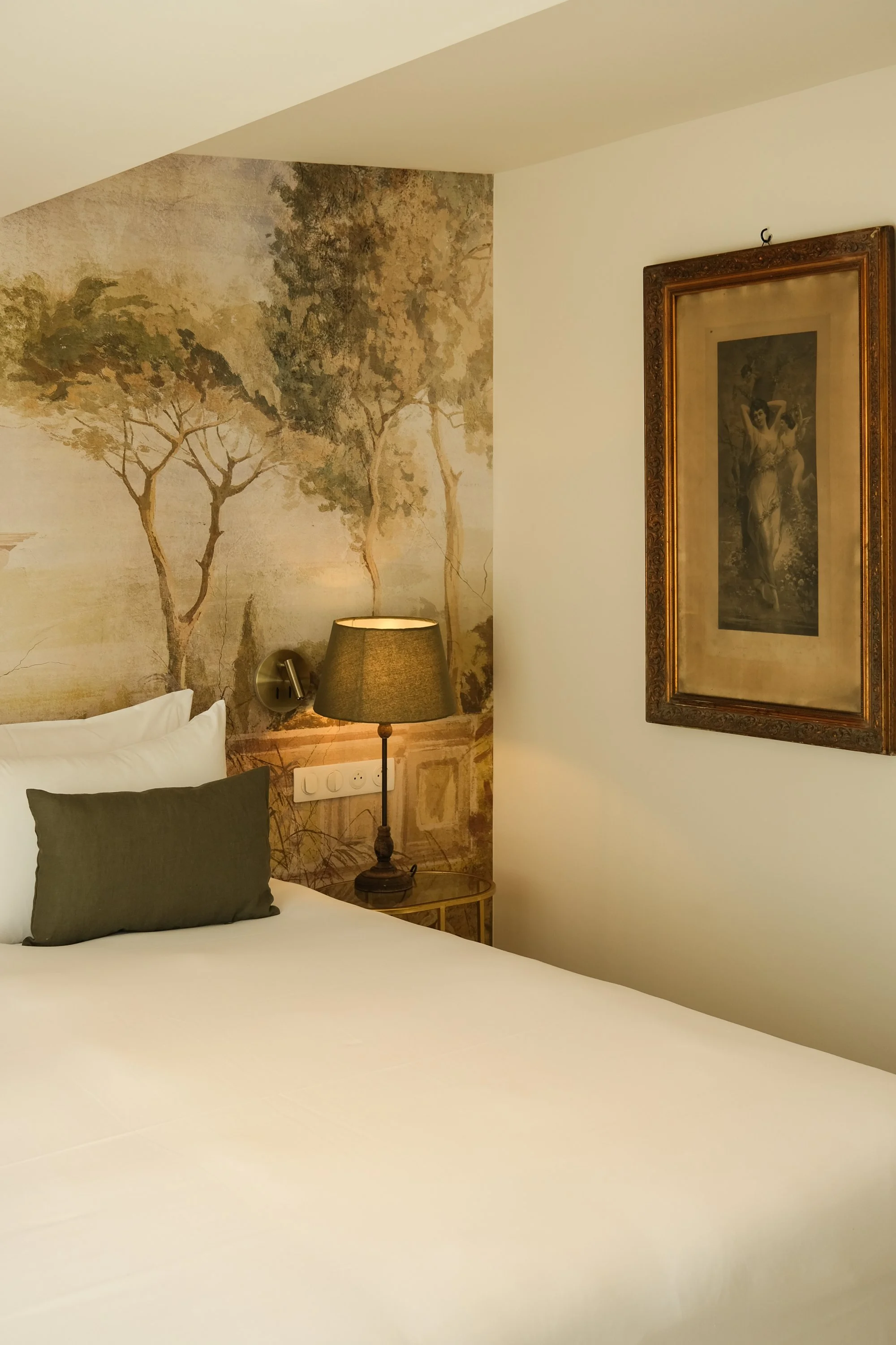 Lit dans une chambre avec une tête de lit blanche, une lampe de chevet, un tableau encadré sur le mur et un mur avec une fresque de paysage avec des arbres.