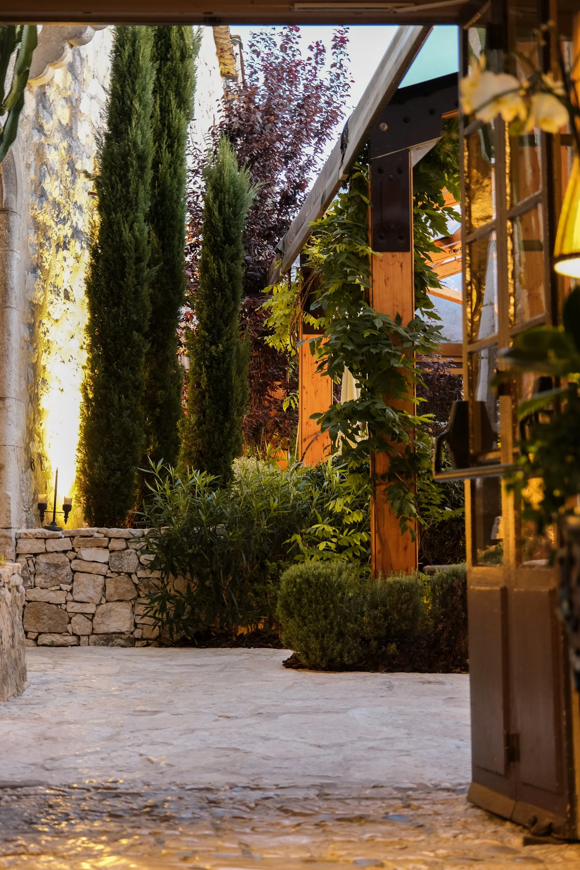 Un jardin avec des plantes vertes et un chemin en pierre, entouré de murs en pierre et d'une structure en bois, avec un éclairage chaud, vu à travers une porte en partie ouverte.
