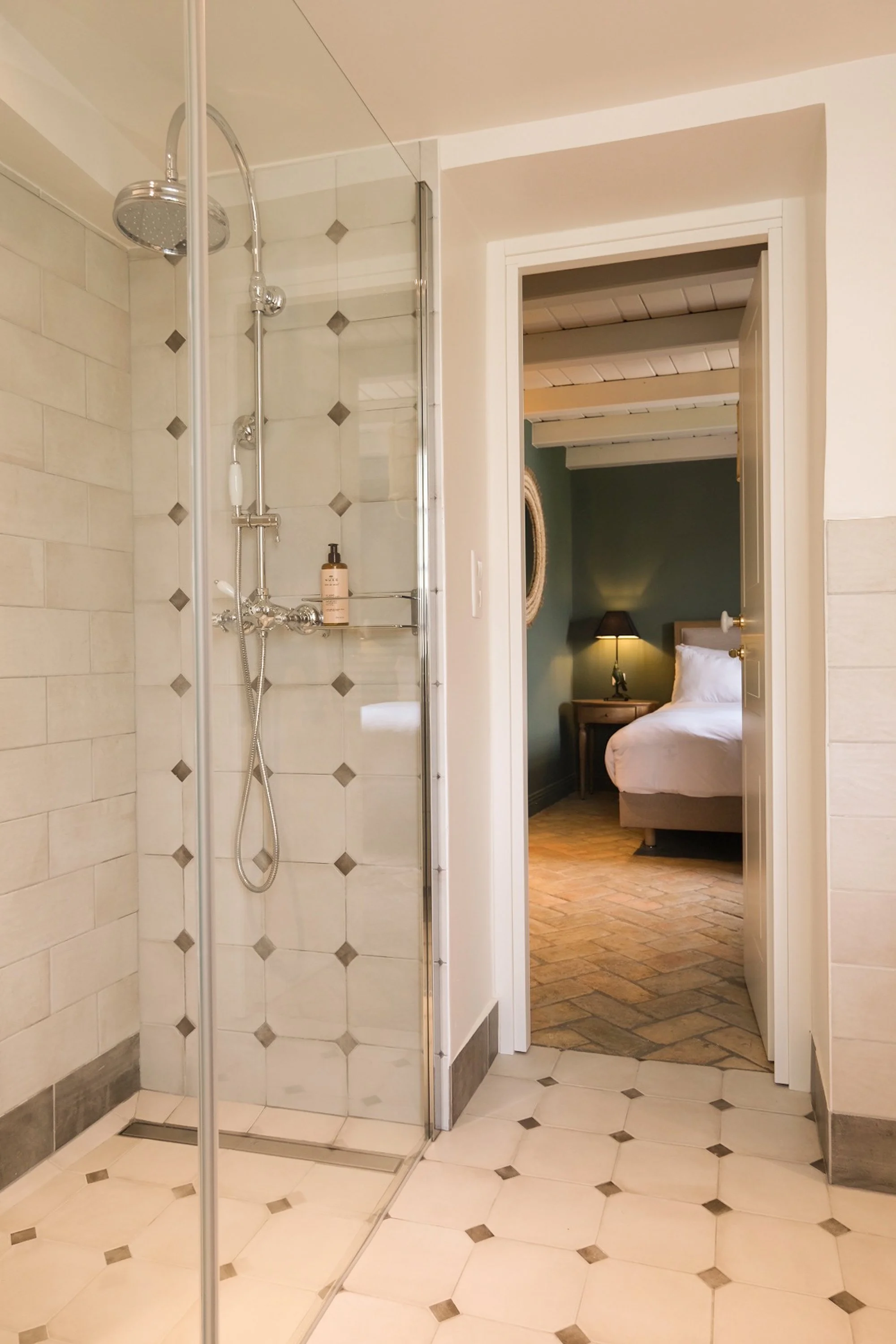 Salle de bain avec douche en verre, vue sur une chambre avec lit et chevet, lampe