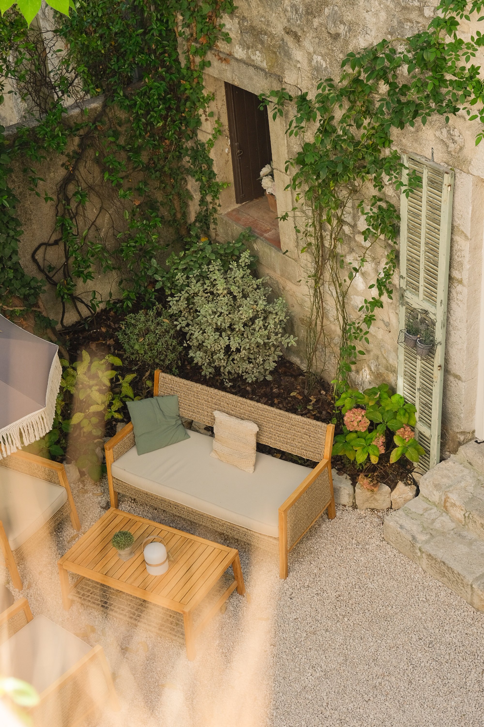 Un petit patio avec un banc en bois, une table basse en bois, un parasol, des plantes grimpantes et des fleurs, entouré d'un mur en pierre et d'une fenêtre avec volets en bois.