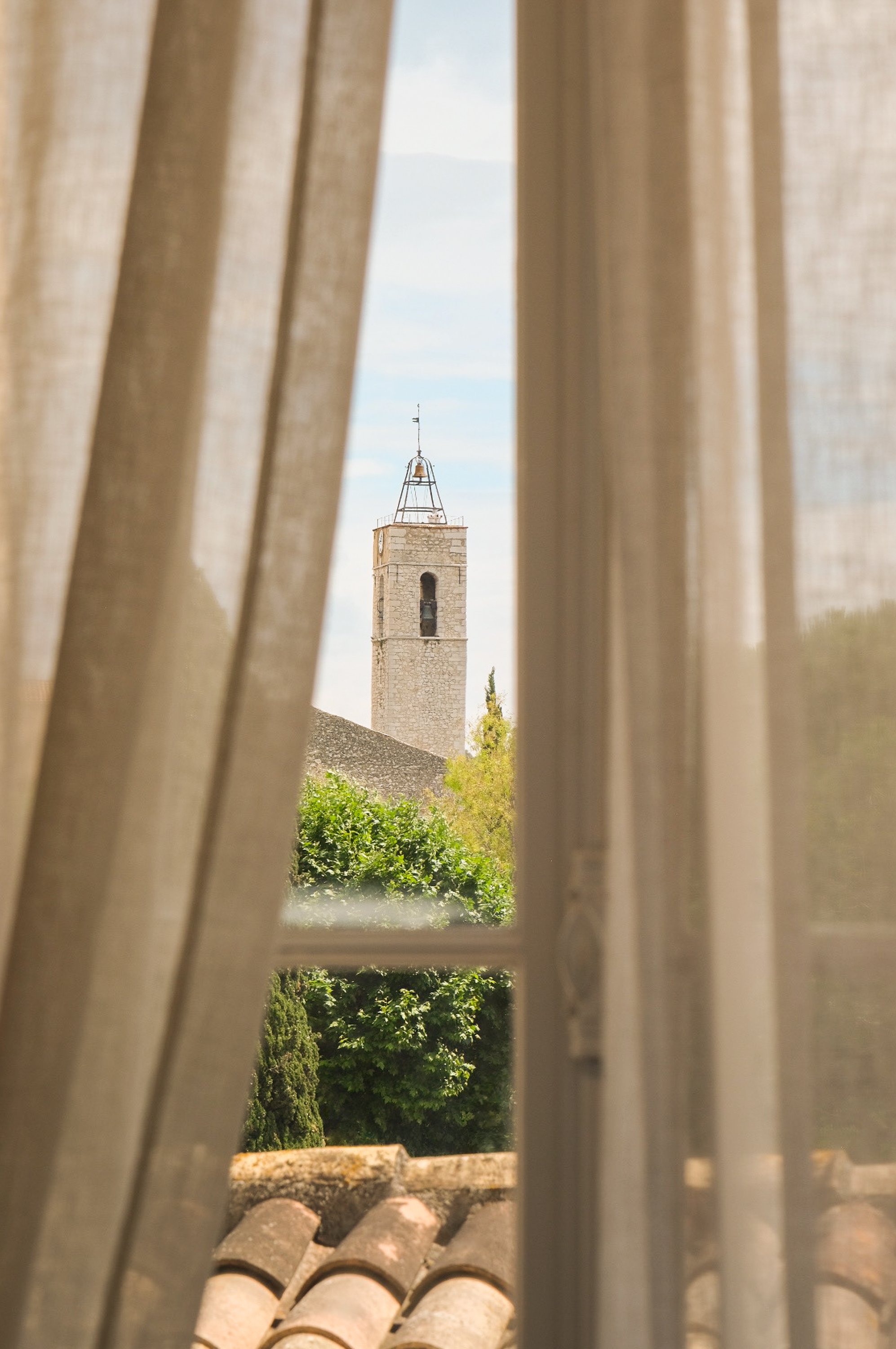 Vue sur le clocher de l'église de Saint Paul de Vence à travers une fenêtre avec rideaux ouverts, arbres verts en dessous.