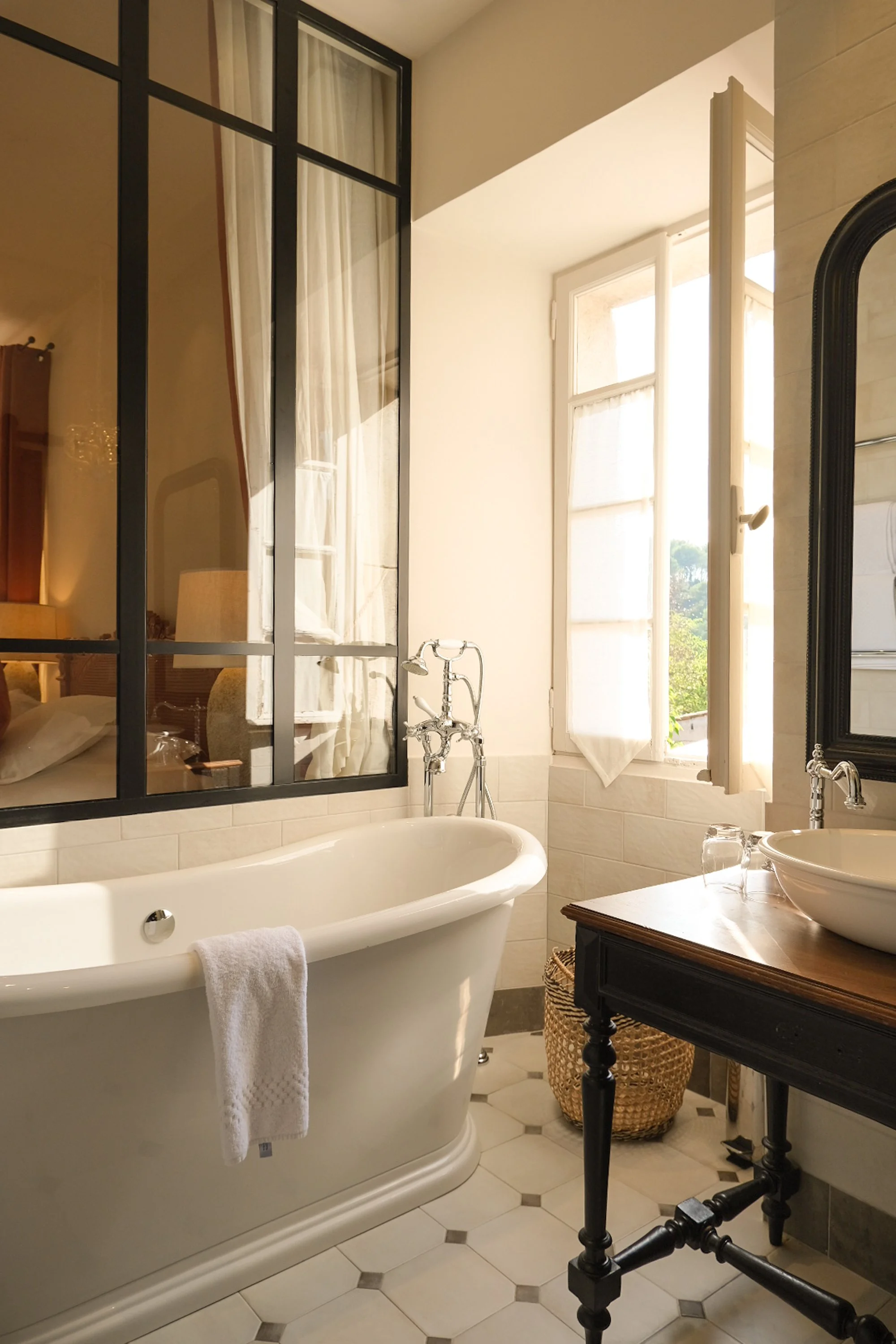 Salle de bain avec une baignoire élégante, un lavabo sur un meuble en bois sombre, une fenêtre laissant entrer la lumière naturelle, et un miroir noir. Un panier en osier est placé contre le mur.