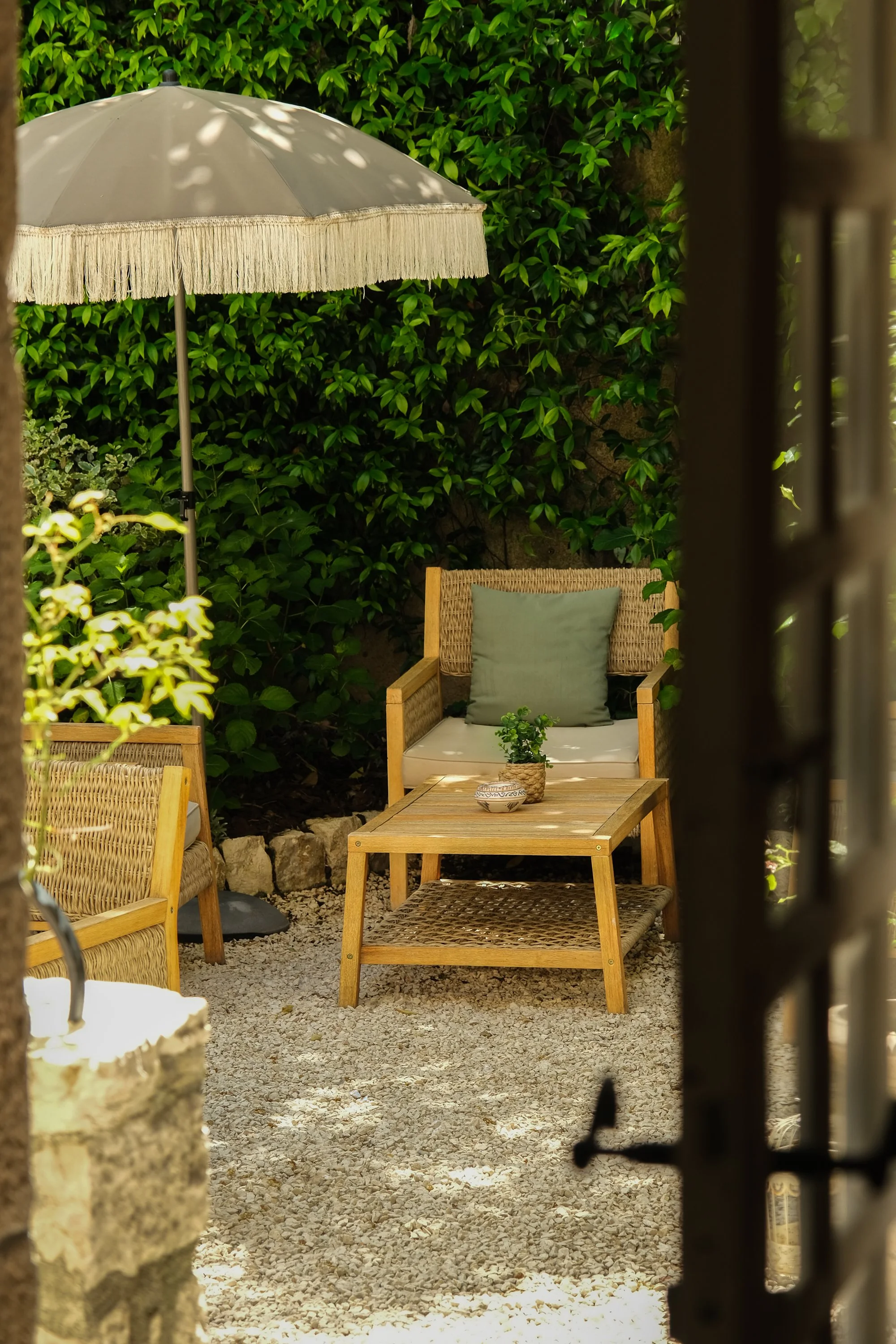 Un coin de jardin avec un fauteuil en bois, une table, une plante en pot, un parasol blanc avec bord frangé, entouré de végétation verte.