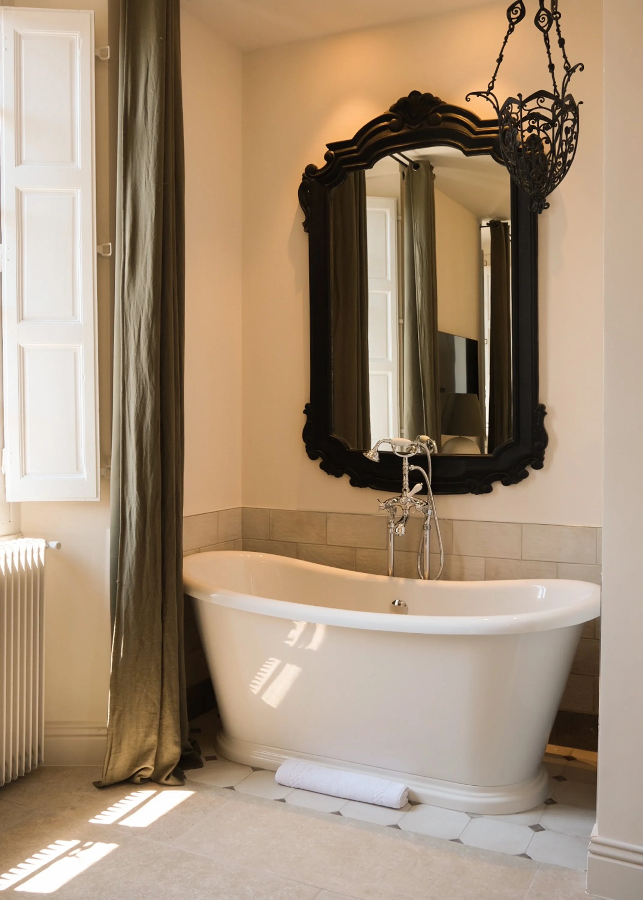 Baignoire blanche dans une salle de bain avec grand miroir noir, chandelier en fer forgé, rideaux vert foncé, fenêtres avec volets, et tapis blanc.