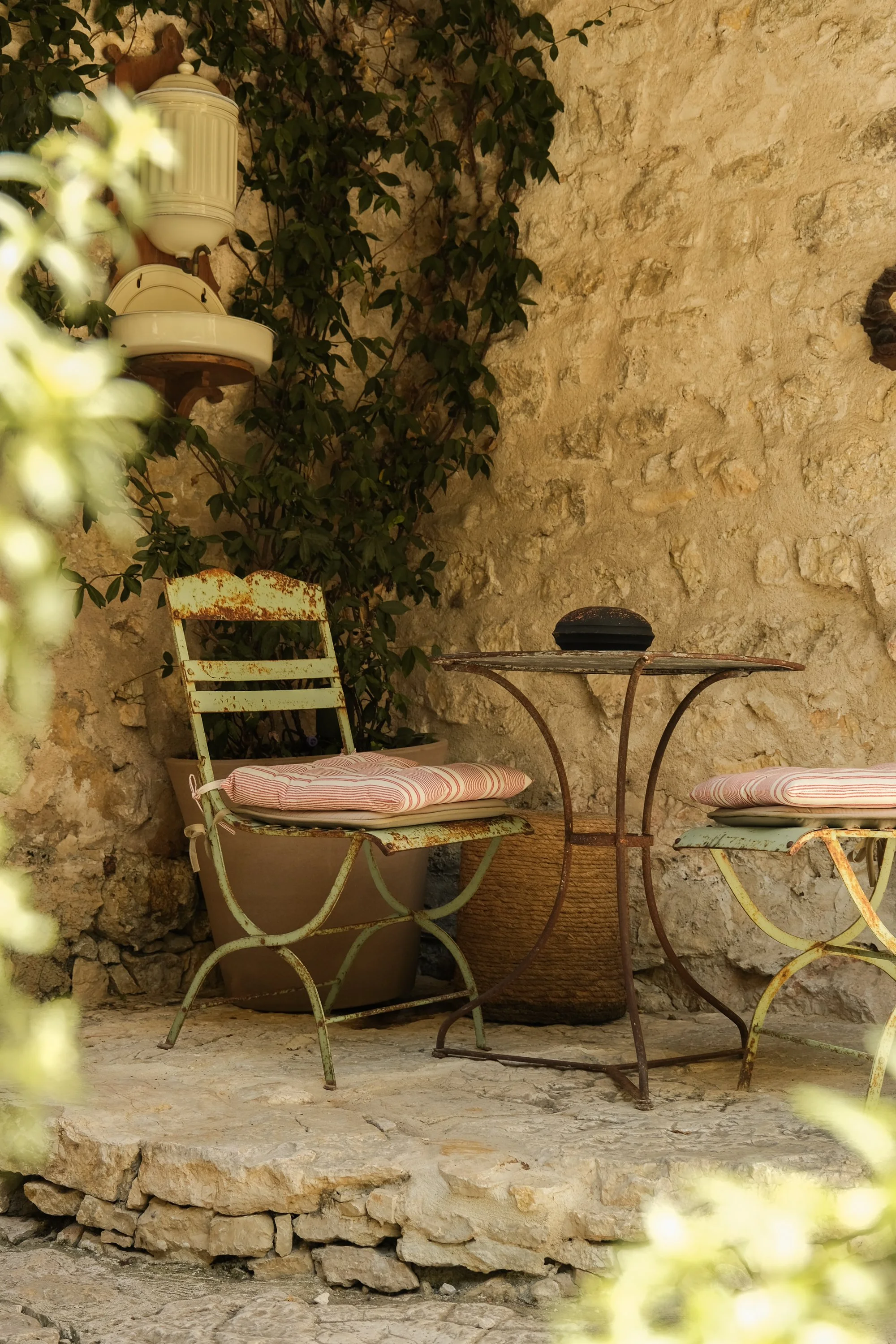 Petite terrasse avec deux chaises en métal peint vert, une petite table en métal rouillé, un grand pot en terre cuite, un mur en pierre, et des plantes en pot avec une toile de fond de plantes grimpantes.