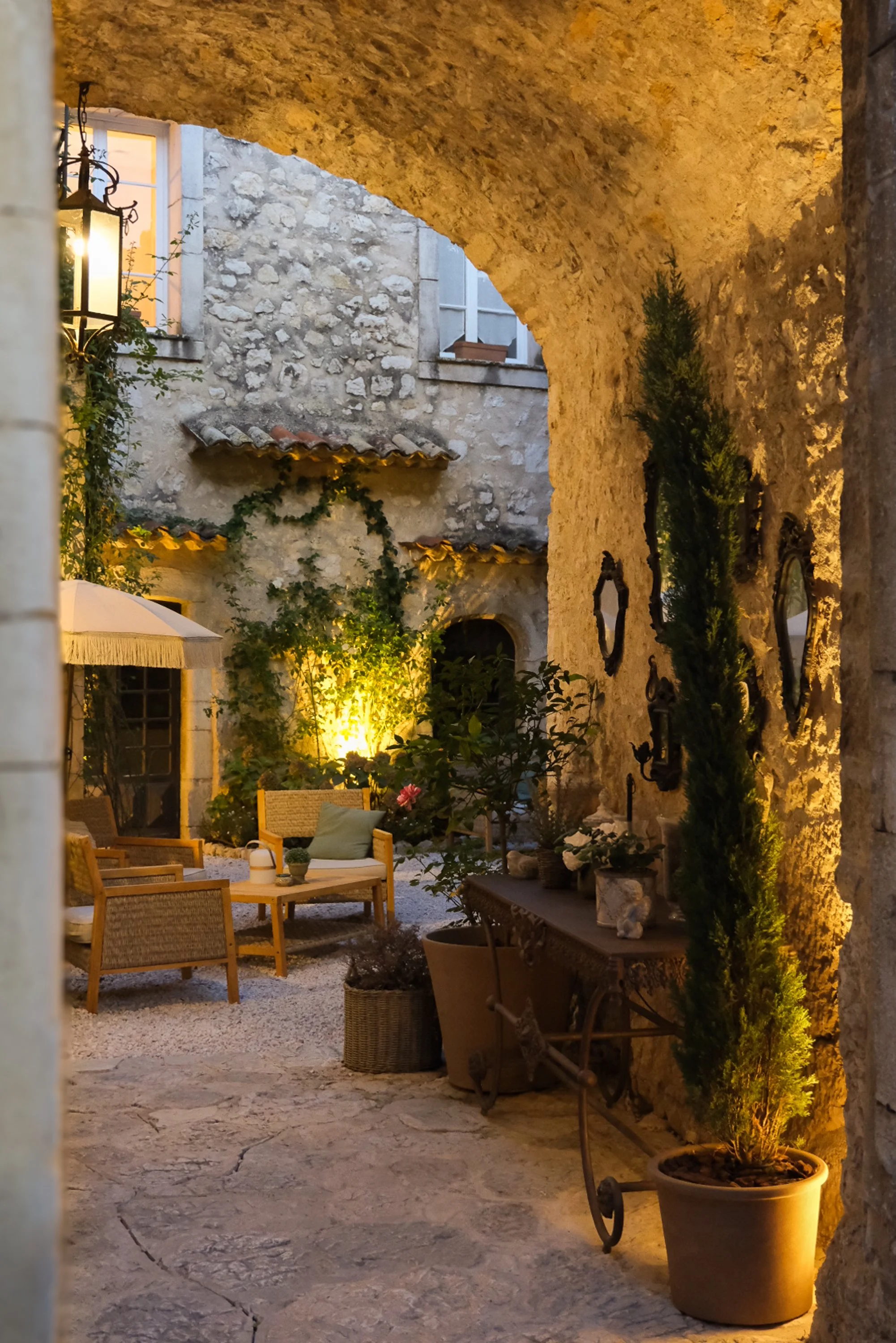 Cour intérieure avec mobilier en rotin, plantes en pot, murs en pierre, éclairage chaleureux et fenêtre en arrière-plan.