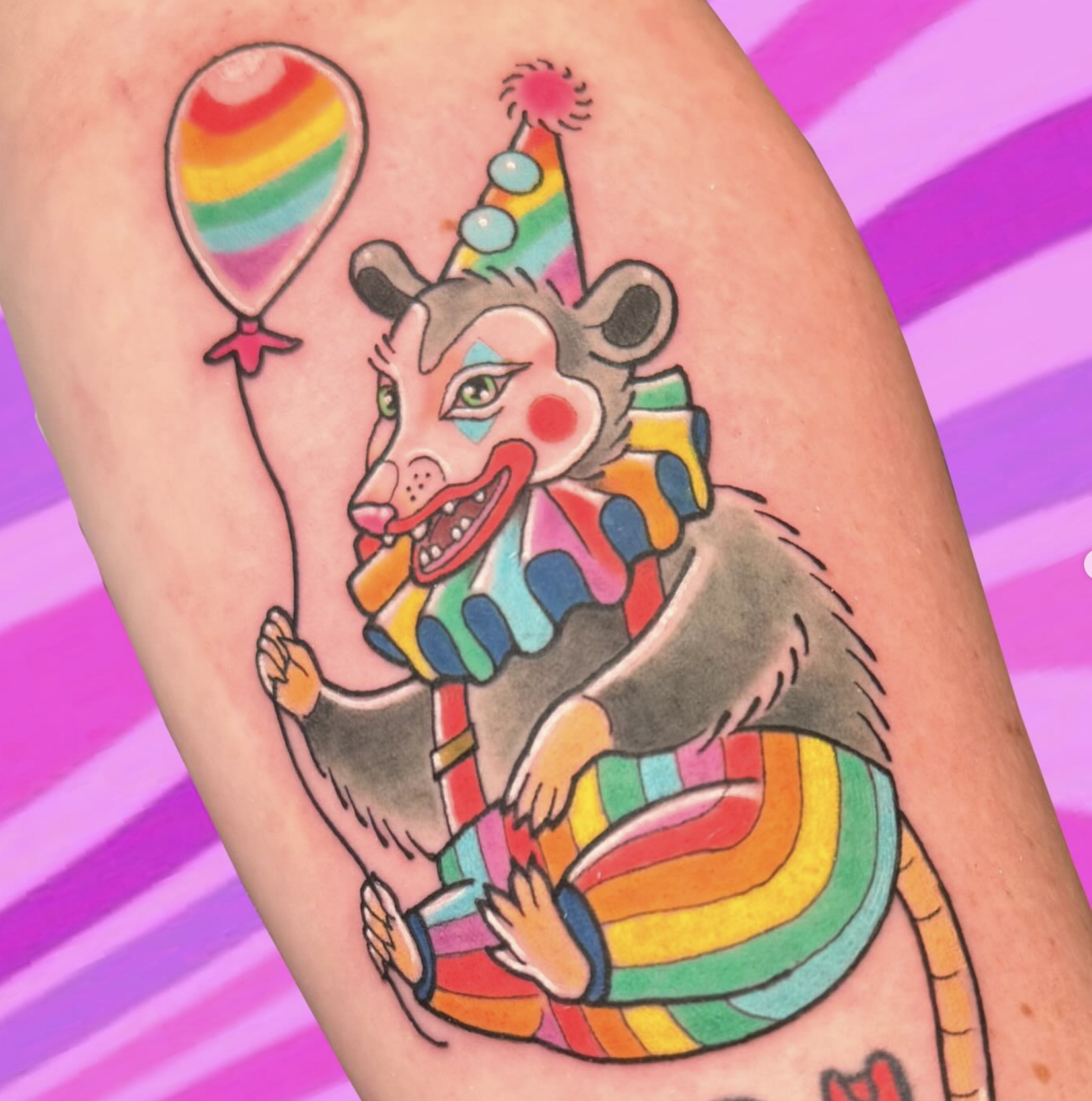 Tattoo of a colorful, whimsical clown rat holding a rainbow balloon, with a rainbow tail and rainbow-striped clothing.