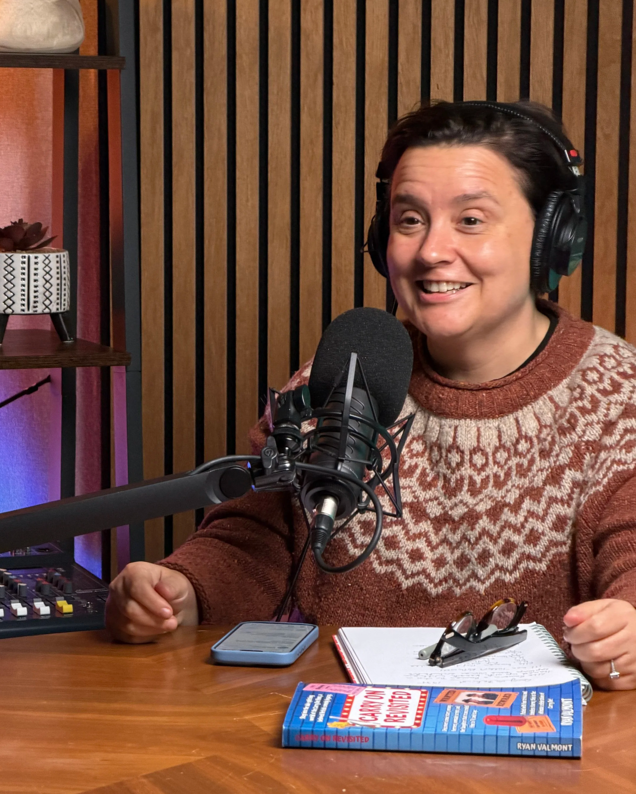 A person with dark hair wearing a patterned sweater and headphones sitting in a recording studio, speaking into a microphone with a closed book, a notebook, and glasses on the table in front of them.