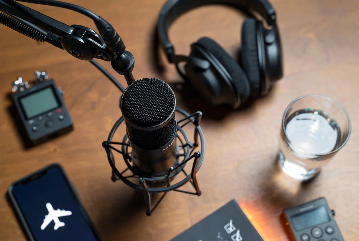 Podcast recording setup on a wooden desk: black condenser microphone on boom arm and shock mount, over-ear headphones, glass of water, portable audio recorder, smartphone with airplane icon, and checklist.