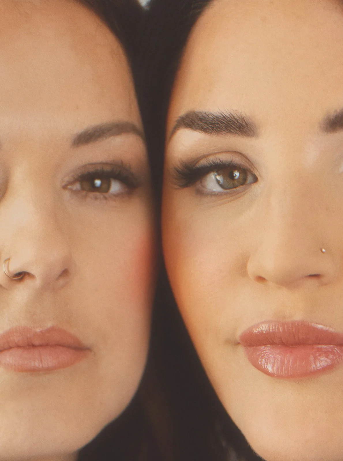 Close-up of two women's faces with makeup, touching cheeks together, showing their eyes, eyebrows, and lips.