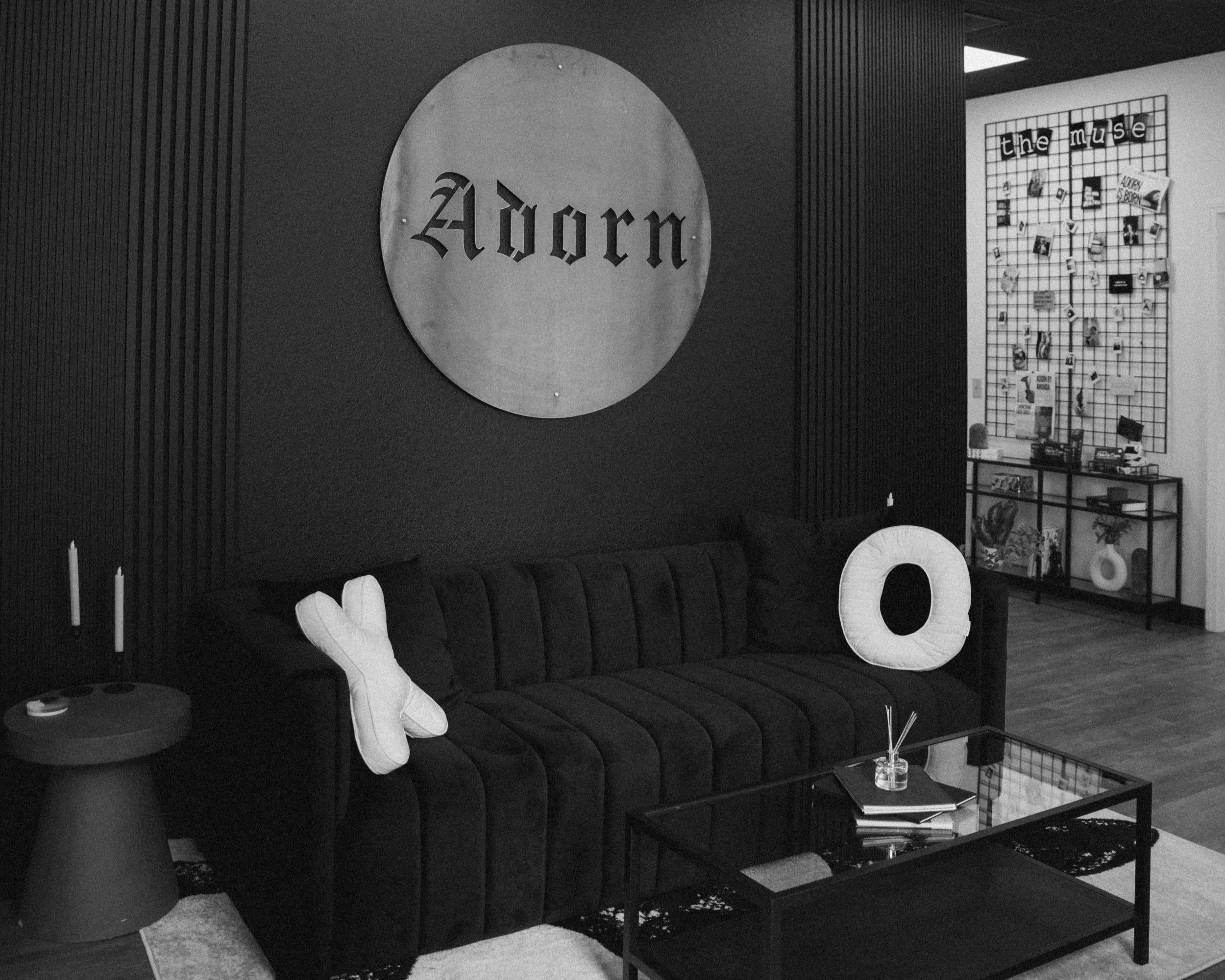 A black sofa with two decorative pillows, one shaped like a bow and the other a round O shape, in a modern living room. Behind the sofa, a large round sign with the word 'Adorn' hangs on the wall. There is a small side table with candles and a black coffee table with books and decor items in front of the sofa. In the background, a wall display with the words 'the muse' and various photos and items is visible.