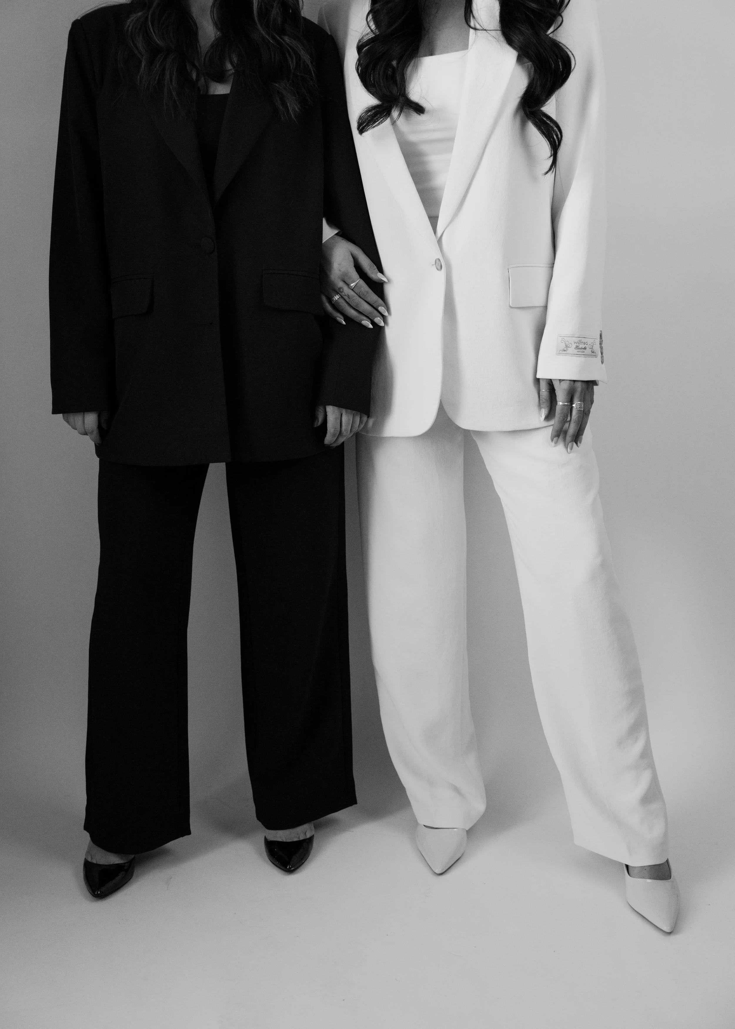 Two women dressed in formal suits, one in black and one in white, standing close together with their arms linked.