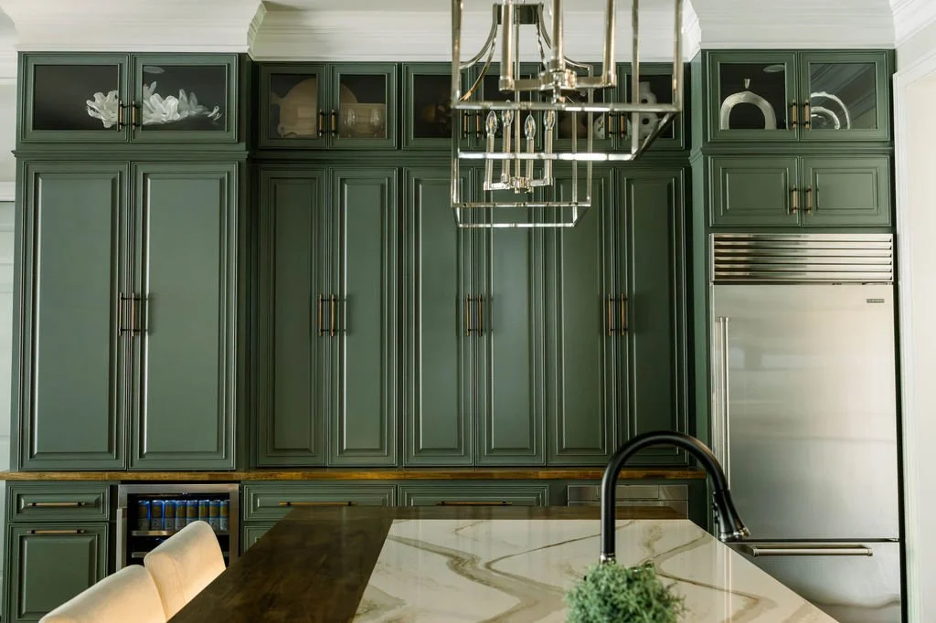 Kitchen with dark green cabinetry, stainless steel refrigerator, and modern chandelier overhead, with a beige chair and a marble countertop island.
