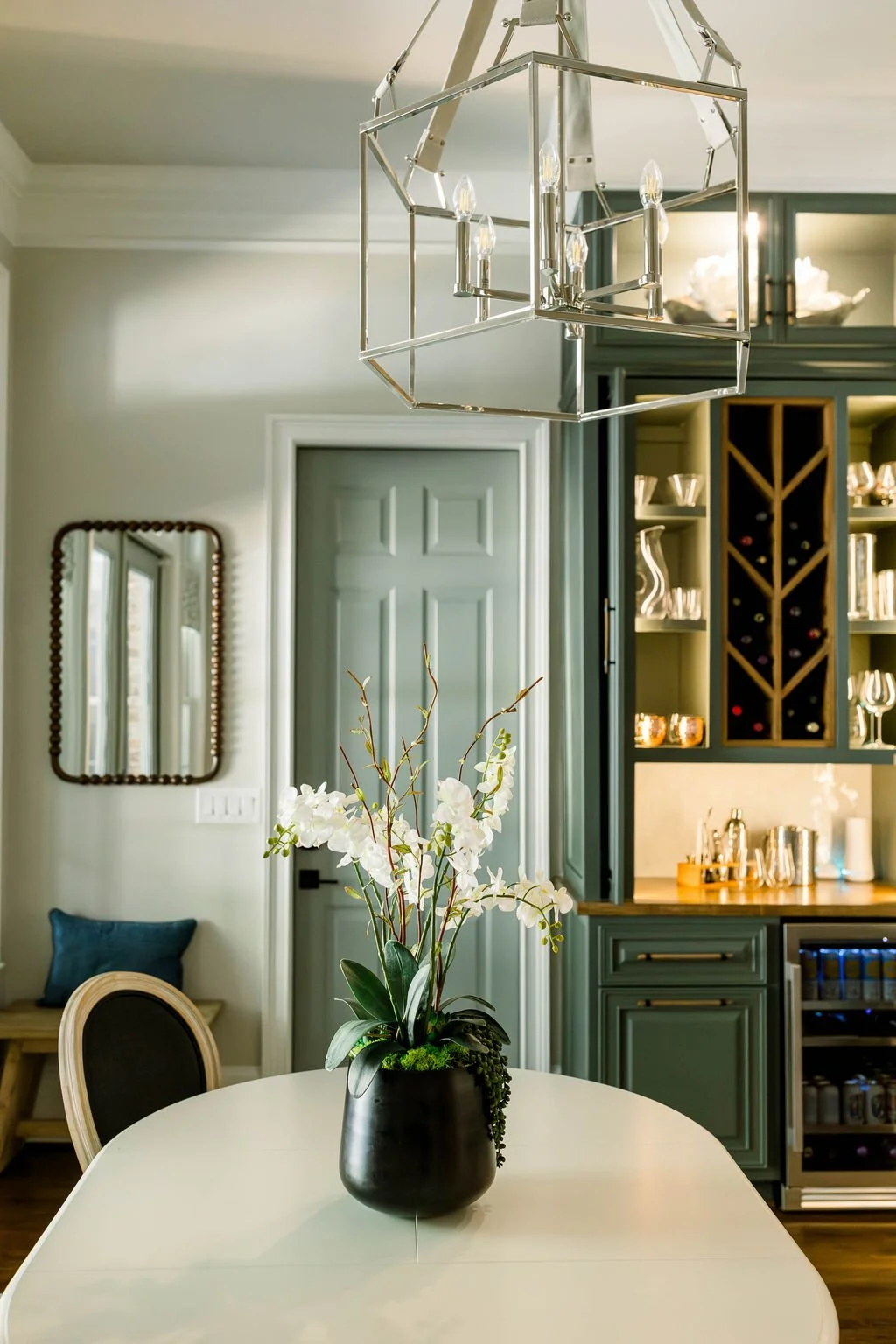 Interior of a modern dining room with a white table, a black vase with white orchids, a teal cabinet, and a geometric chandelier.