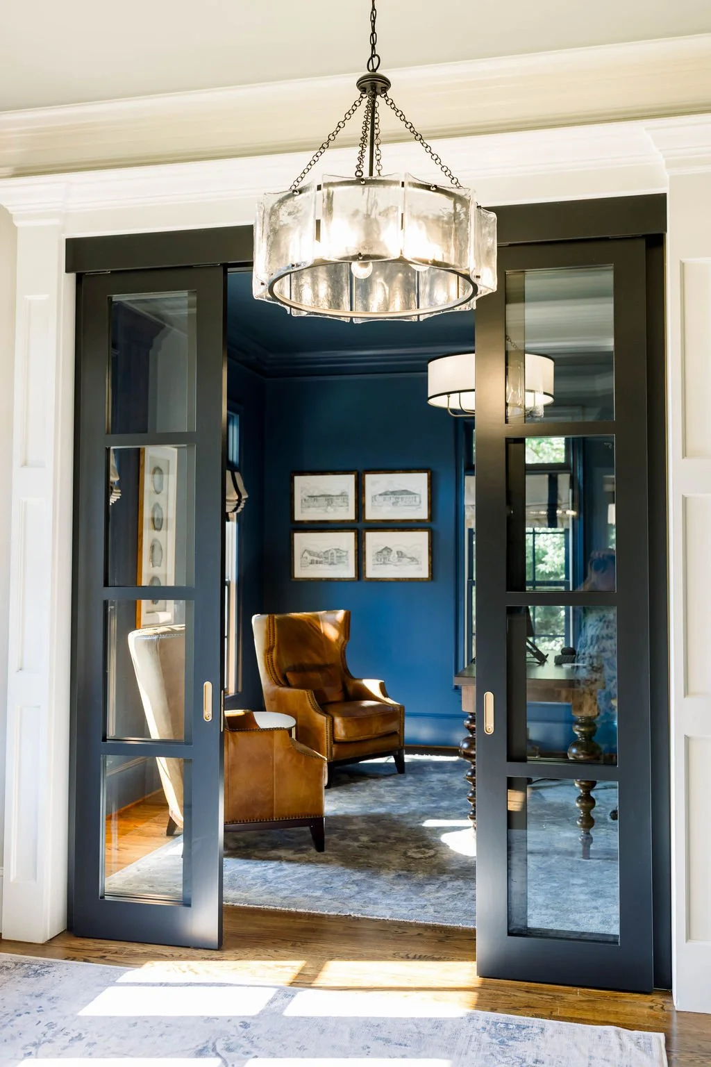 View through black sliding glass doors into a blue sitting room with leather armchair, framed wall art, and a window with a shade.