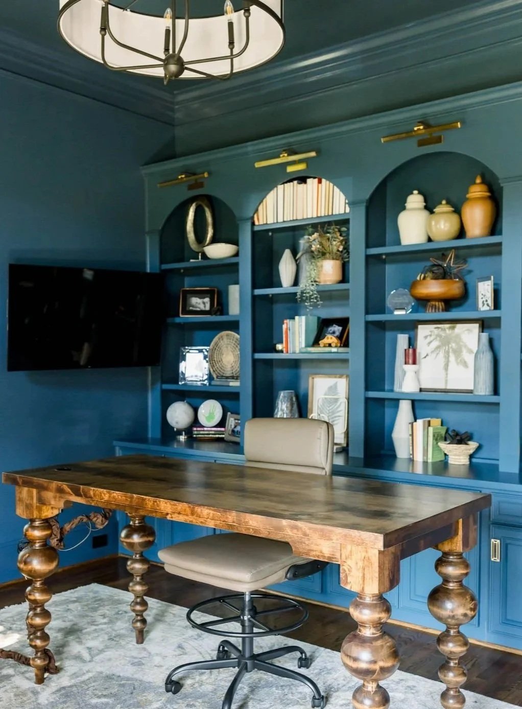 A home office with a large wooden desk and an office chair in front of a blue built-in bookshelf filled with decorative items, books, and framed pictures. A flat-screen TV is mounted on the blue wall to the left of the shelves. A modern chandelier ha