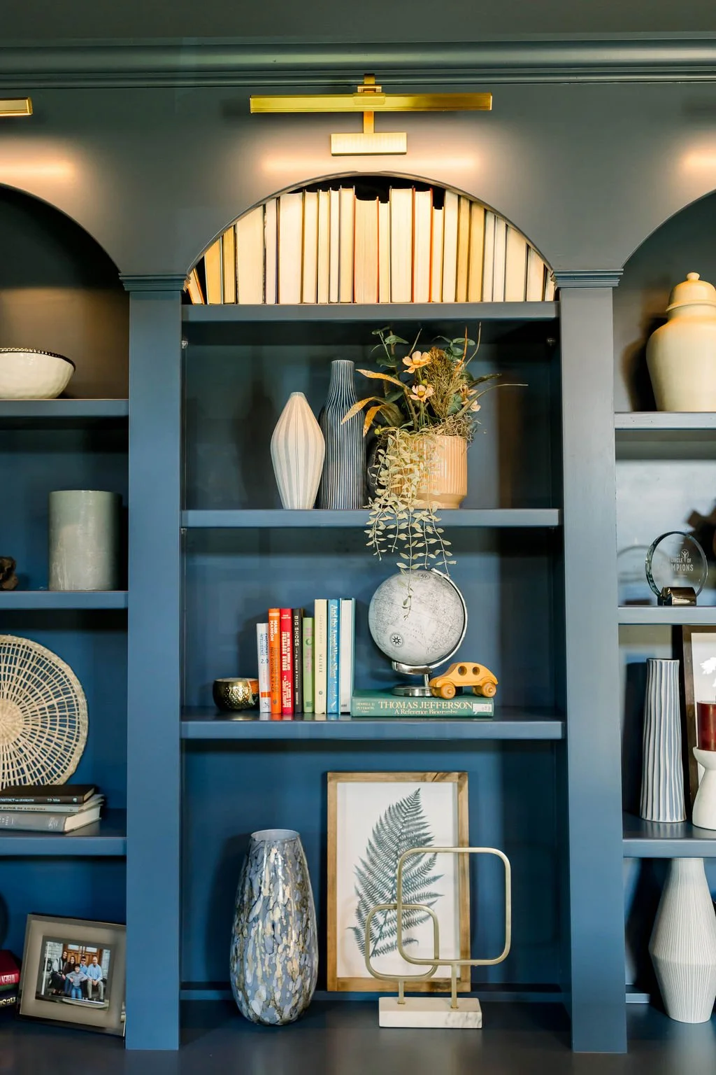 Blue built-in bookshelf with decorative vases, books, framed art, and plants, illuminated by warm lights.