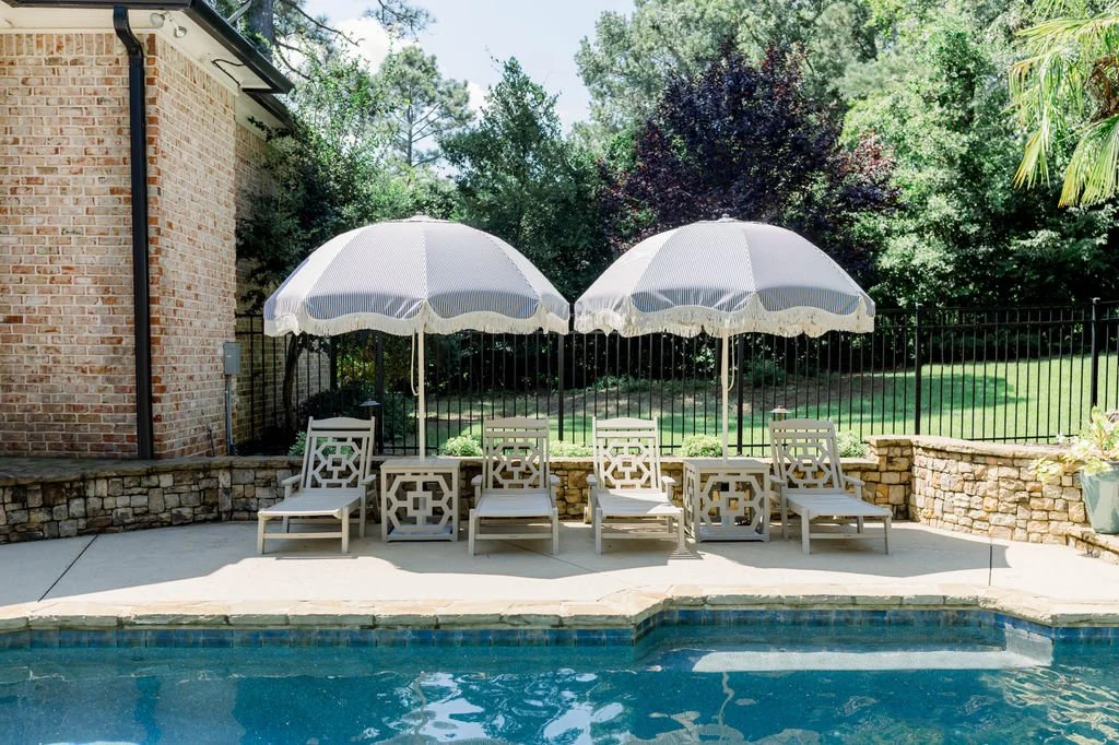 Poolside scene with four white lounge chairs, two white umbrellas, a stone wall, and a green backyard with trees and grass.