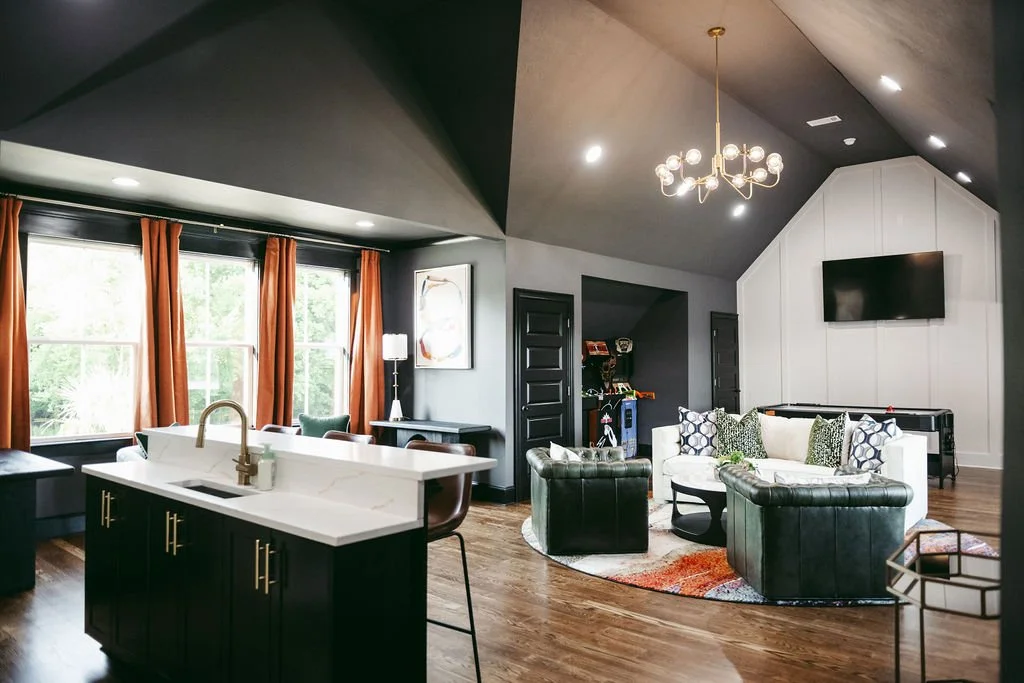 Open-concept living room and kitchen area with dark ceiling, large windows with orange curtains, white walls, black doors, modern chandelier, flat-screen TV, black leather chairs, white sofa with patterned pillows, and a small round rug on wood flooring.