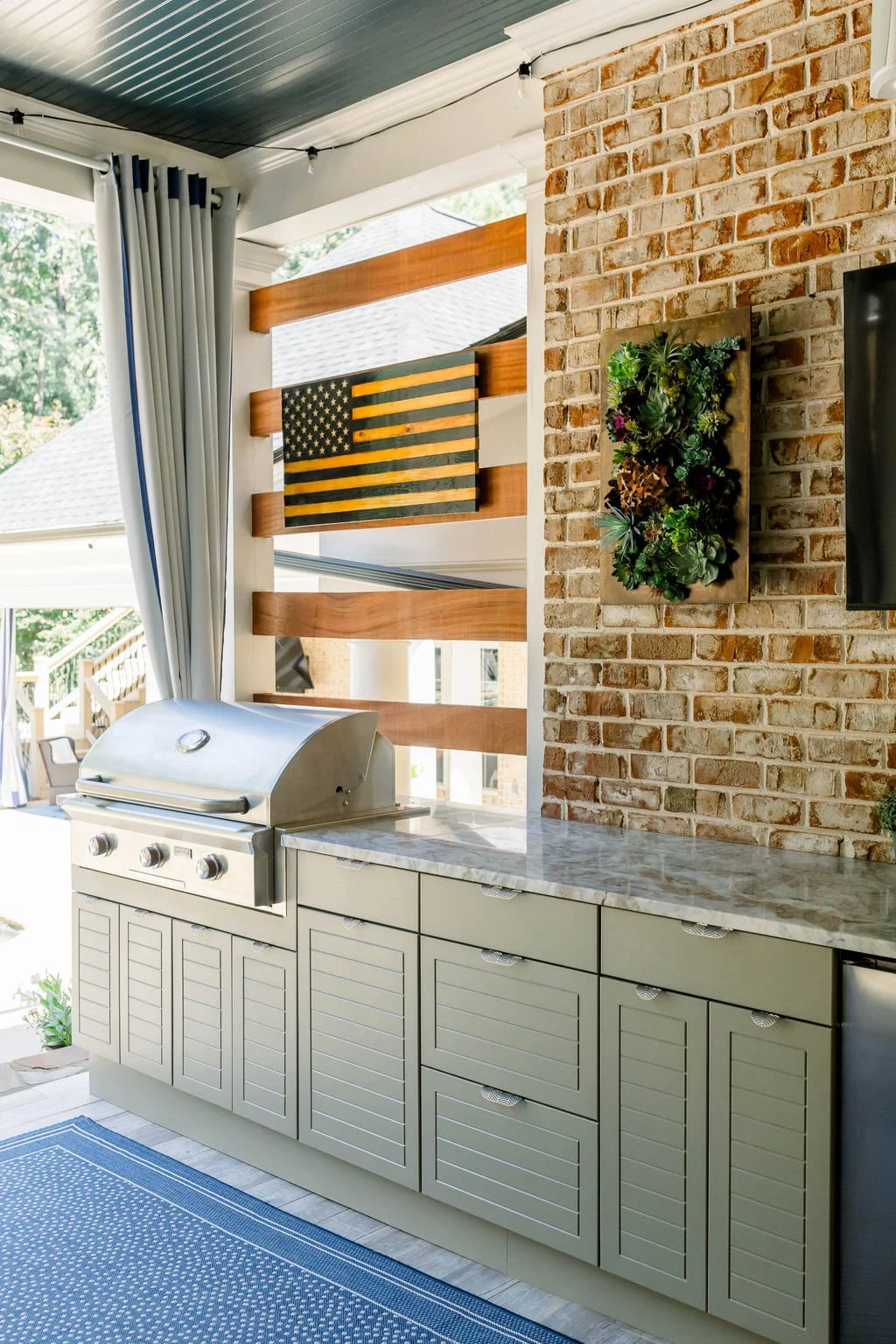 Outdoor kitchen area with grill, green cabinetry with shutters, brick wall with a vertical garden, American flag art on the wall, string lights, and large windows with curtains.