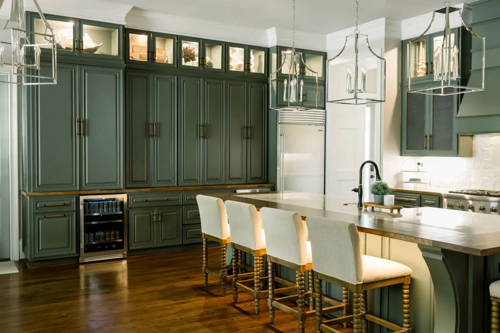 Green kitchen with cabinetry, a wooden island with white upholstered chairs, and modern chandeliers.