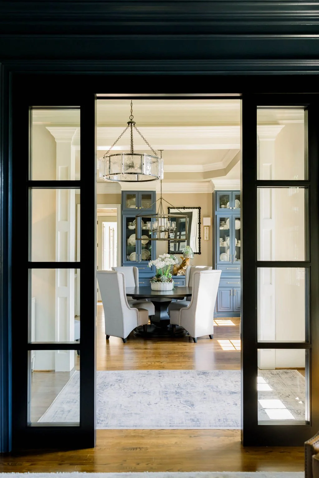 View through a black-framed glass door into a dining room with a round table surrounded by white upholstered chairs, blue china cabinets, a chandelier, and a mirror on the wall.