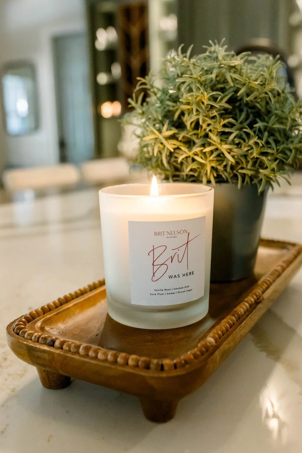 A lit white candle labeled 'Brit' placed on a wooden tray, with a potted green plant behind it, on a table in a cozy indoor setting.