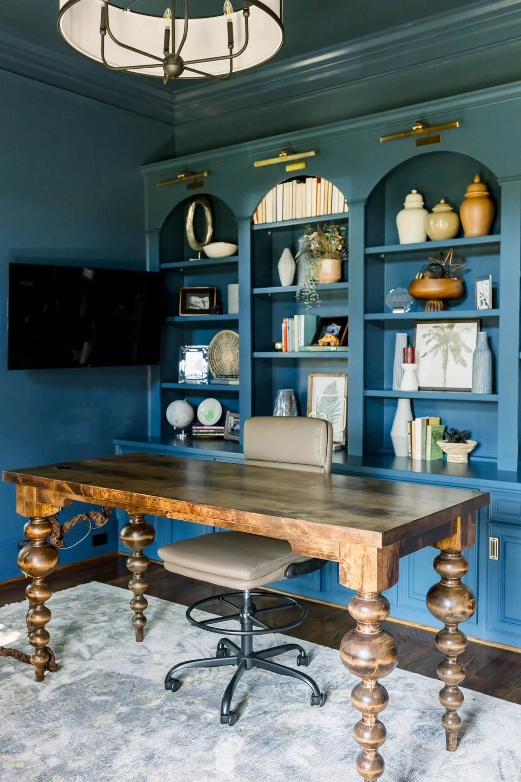 A home office with a large wooden desk and an office chair in front of a blue built-in bookshelf filled with decorative items, books, and framed pictures. A flat-screen TV is mounted on the blue wall to the left of the shelves. A modern chandelier ha