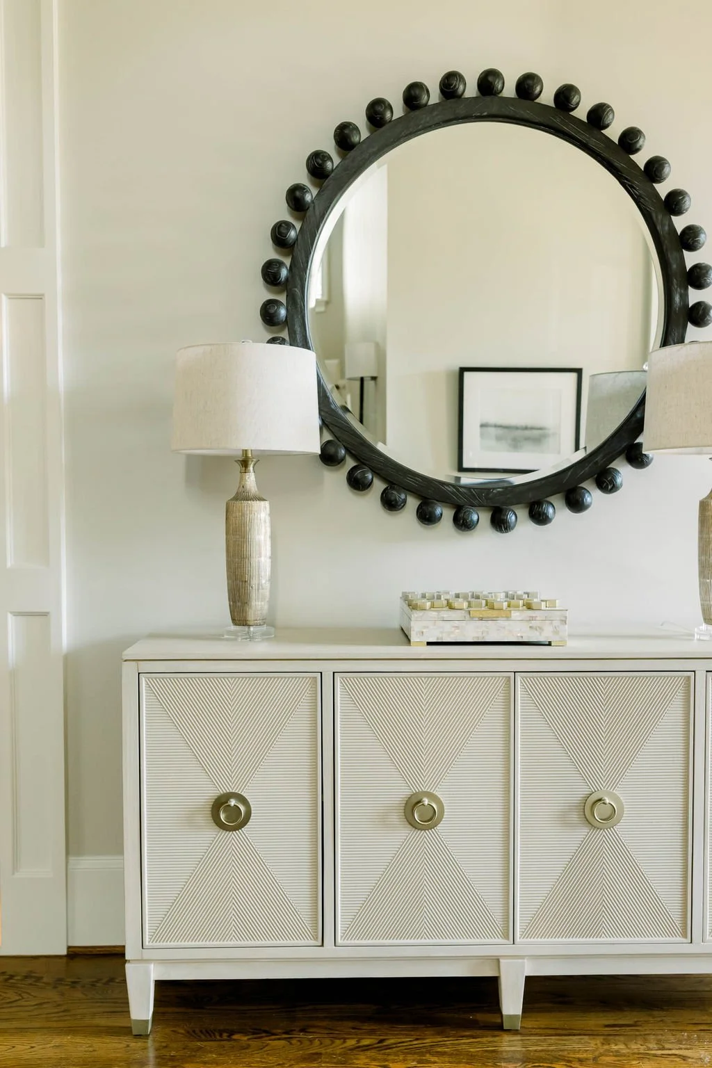 A white sideboard with textured drawer fronts and gold ring pulls, topped with a tall beige lamp and a decorative box, is reflected in a large, round black-framed mirror with ball accents.