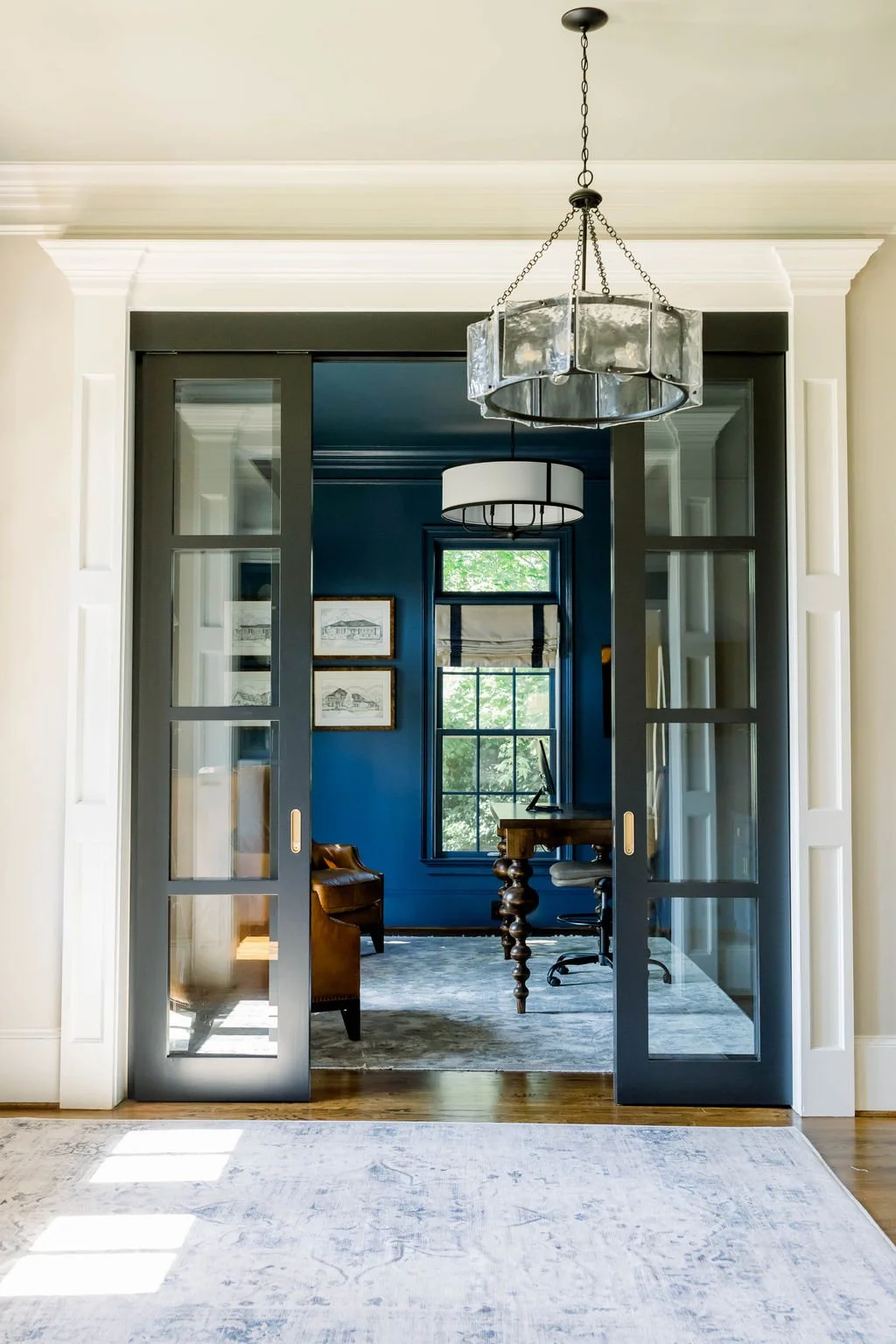 Interior view of a home office with dark blue walls, a wooden desk, a brown leather armchair, framed artwork, and large glass-paneled sliding doors leading to a bright room.