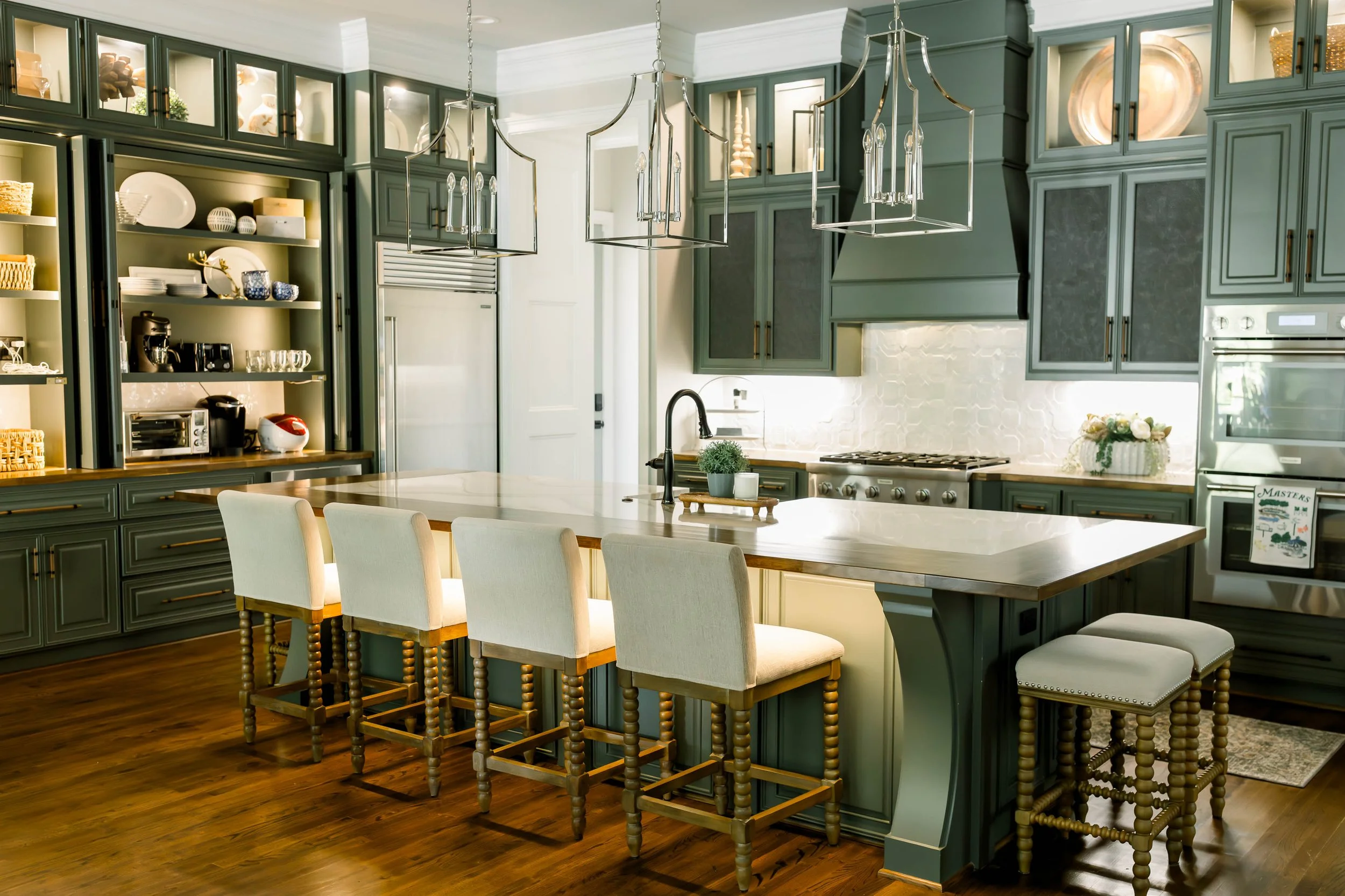 Modern kitchen with dark green cabinetry, a large white island with seating, pendant lights, and stainless steel appliances.