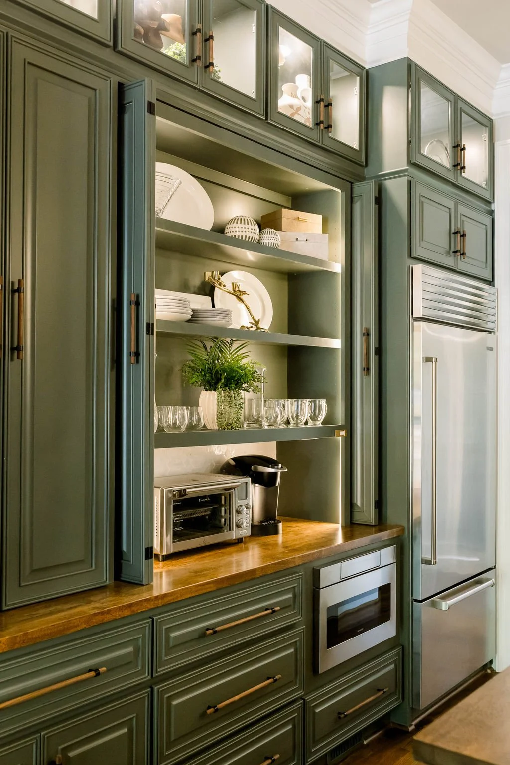 Kitchen cabinet with open shelves displaying dishes, glasses, decorative boxes, and a plant, next to a stainless steel refrigerator.