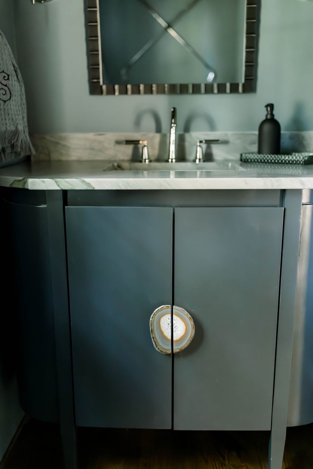 Close-up of a modern kitchen sink with a marble countertop, stainless steel faucet, black soap dispenser, and a soap tray, with reflections visible in the mirror above.