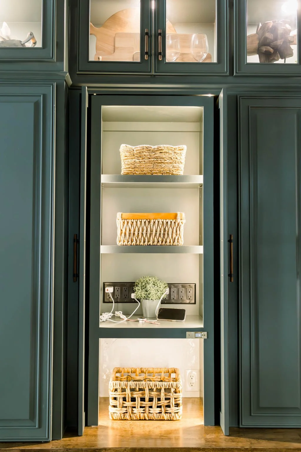 Open built-in cabinet with three shelves containing woven baskets, a small plant, and a tablet. The cabinet is part of a larger dark green storage unit with glass doors on top and closed doors on the sides.