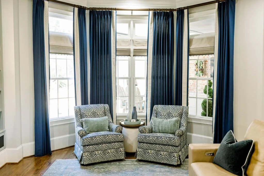 A cozy living room corner with two patterned armchairs and green pillows, a small round side table with vases, large windows with blue curtains, and a glimpse of outdoor trees and brick wall.