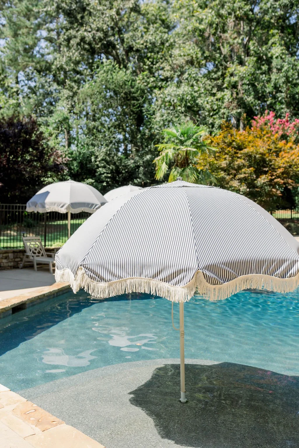 A swimming pool with two large striped umbrellas providing shade, surrounded by trees and outdoor furniture.