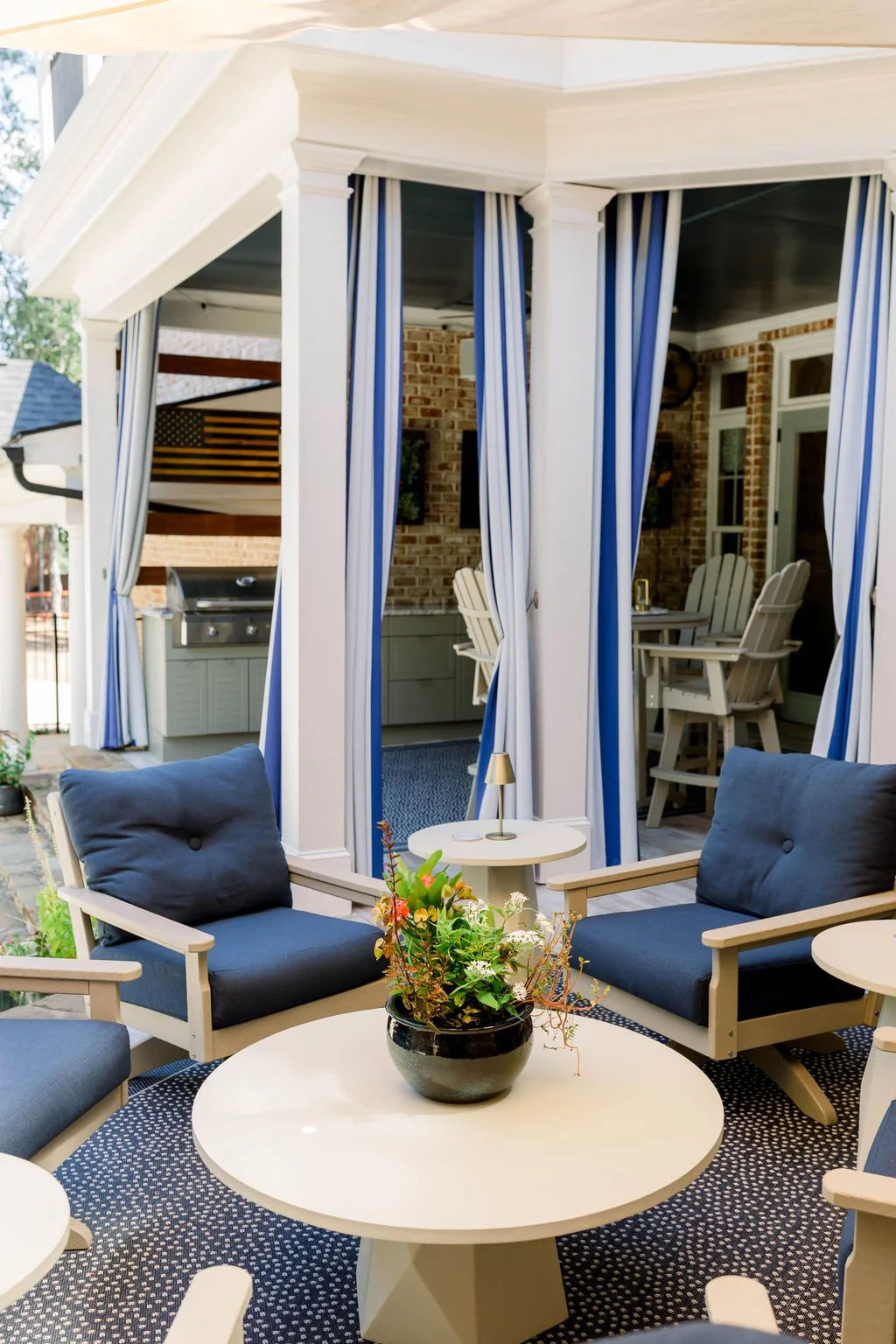 Patio area with navy cushioned chairs, a white table with a flower arrangement, and a covered porch with striped curtains and a dining set in the background.
